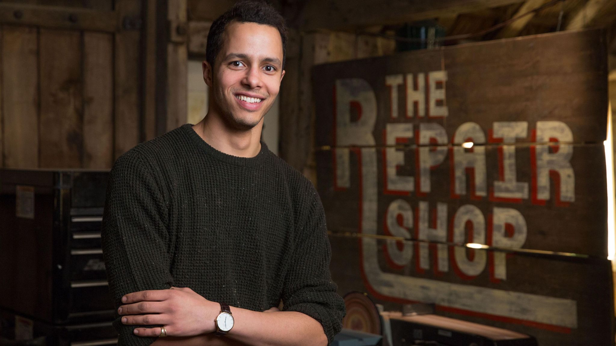 Will Kirk, with short dark hair and wearing a brown sweater, stands in front of the Repair Shop sign