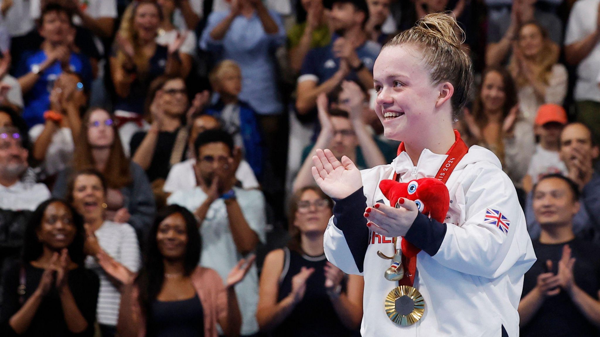 Maisie Summers-Newton wearing a white Team GB sweatshirt smiling and clapping with a gold Paralympic medal around her neck, and a crowd applauding in the background