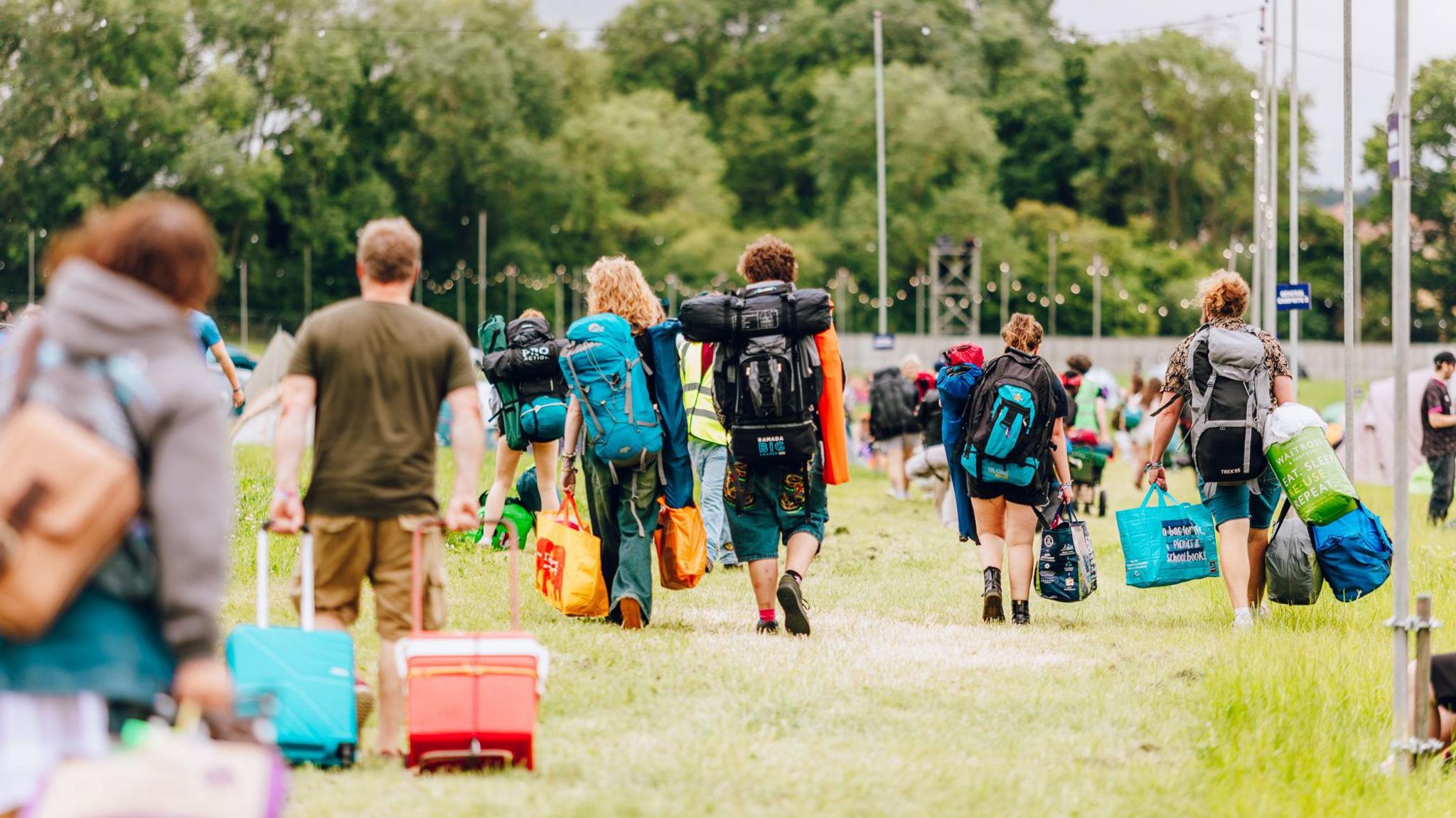 Campers arrive at Truck Festival. Most wear rucksacks and carry bags. A man in front is pulling two wheeled suitcases.