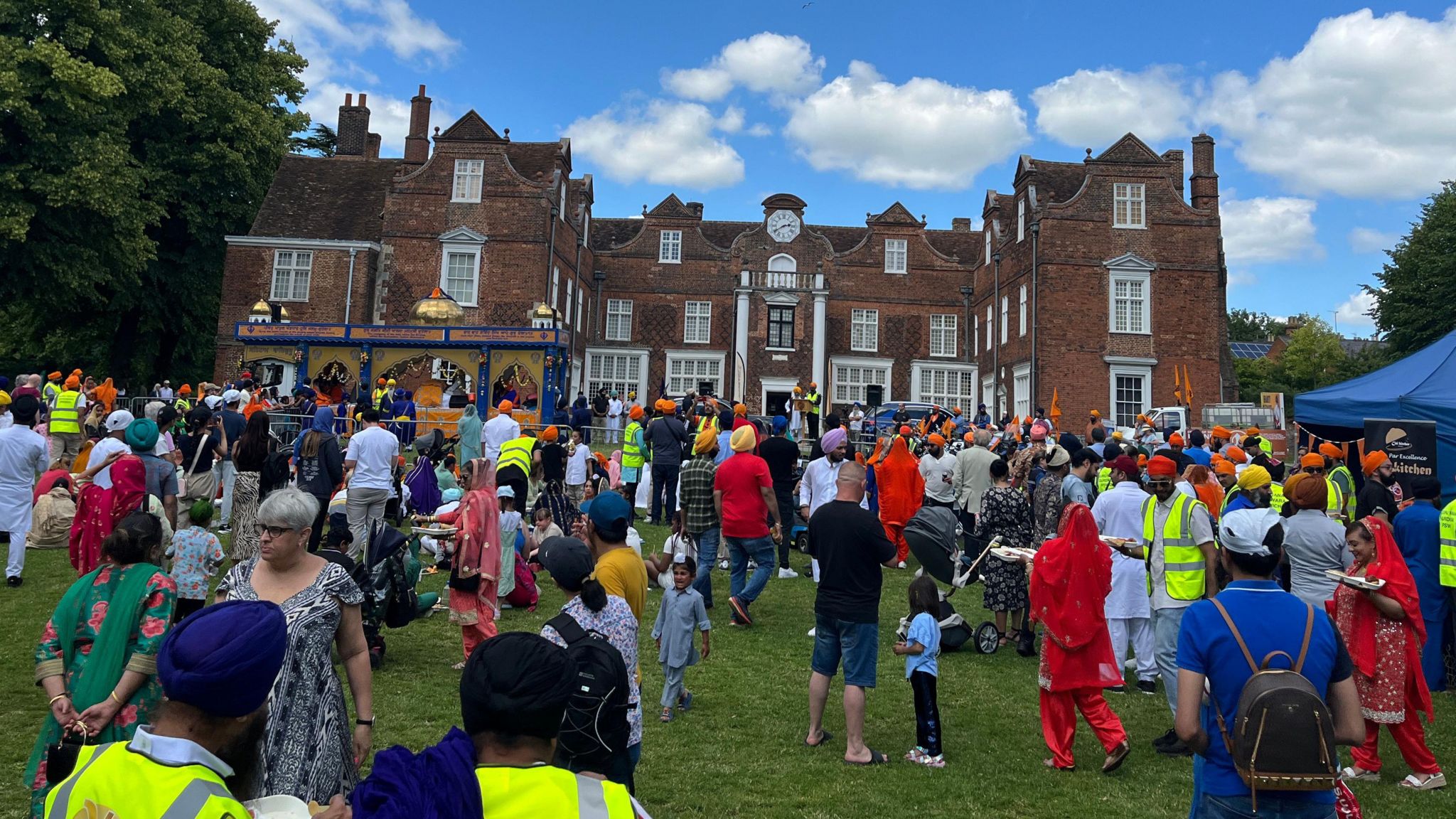 A crowd of people from different nationalities in front of a tudor mansion, with a Sikh float in the background