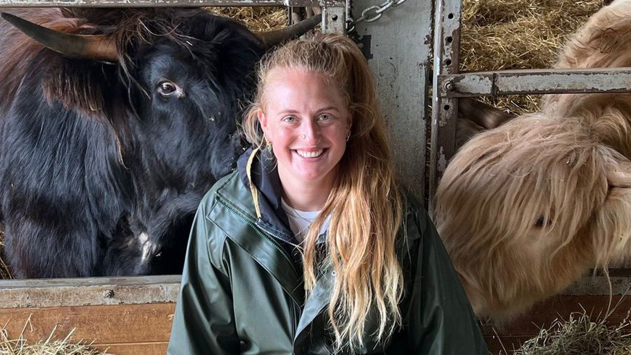 Izzi Rainey - a blonde woman in her early 30s kneels in a barn in front of two of her cattle, one black and one blonde. Izzi s a while woman and her hair, tied in a pony tail, is over her shoulder reaching down to her middle. She wears a dark green rain mac and knee high boots and kneels amongst some hay - both cows are taking a keen interest in Izzi.