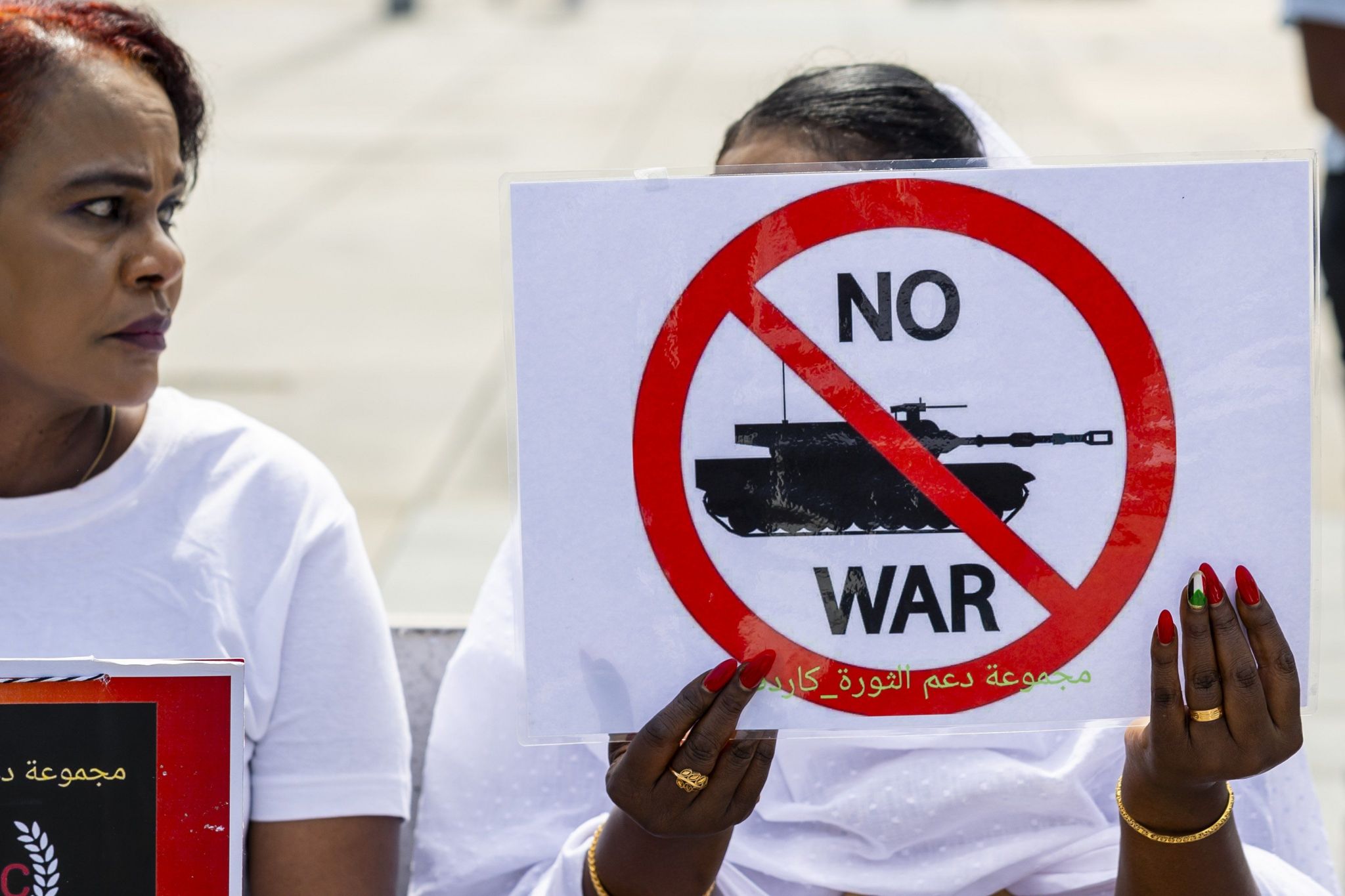 A woman holds up a sign saying 'no war'. It shows an image of a tank under a red stop sign.