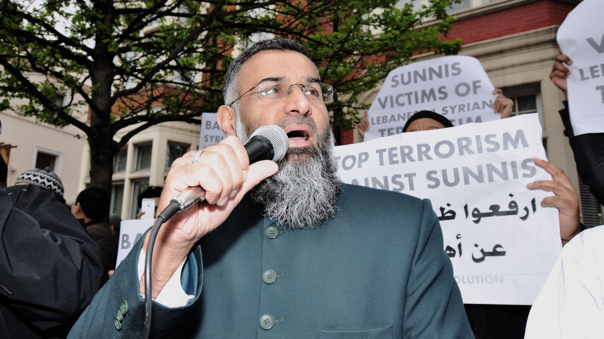 Choudary campaigning in 2014 - he is holding up a mic and is wearing a green jubba - behind him his supporters hold up signs in support