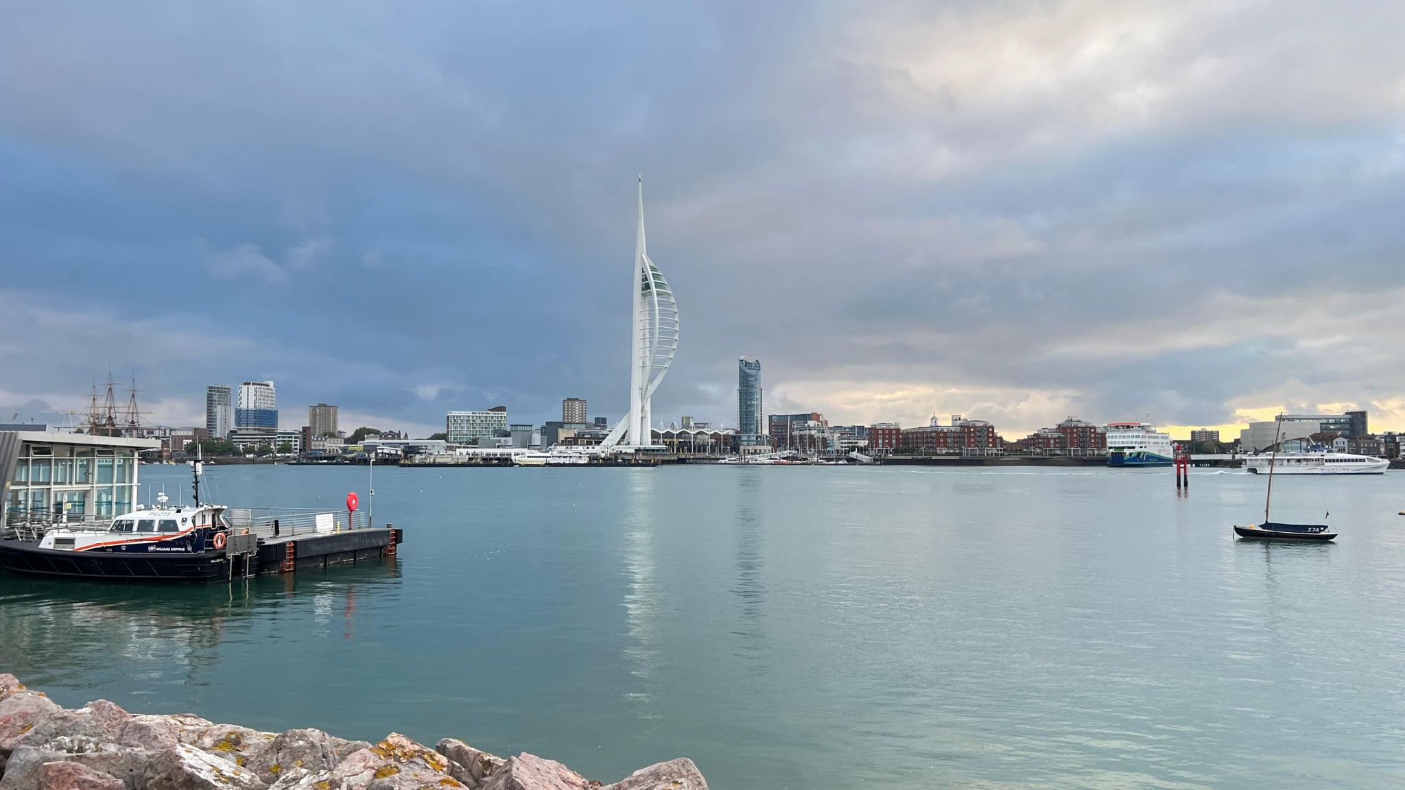 The Spinnaker Tower sits prominently at the back of the photo under clouds and in front of a large area of water that is largely aside from a few small boats