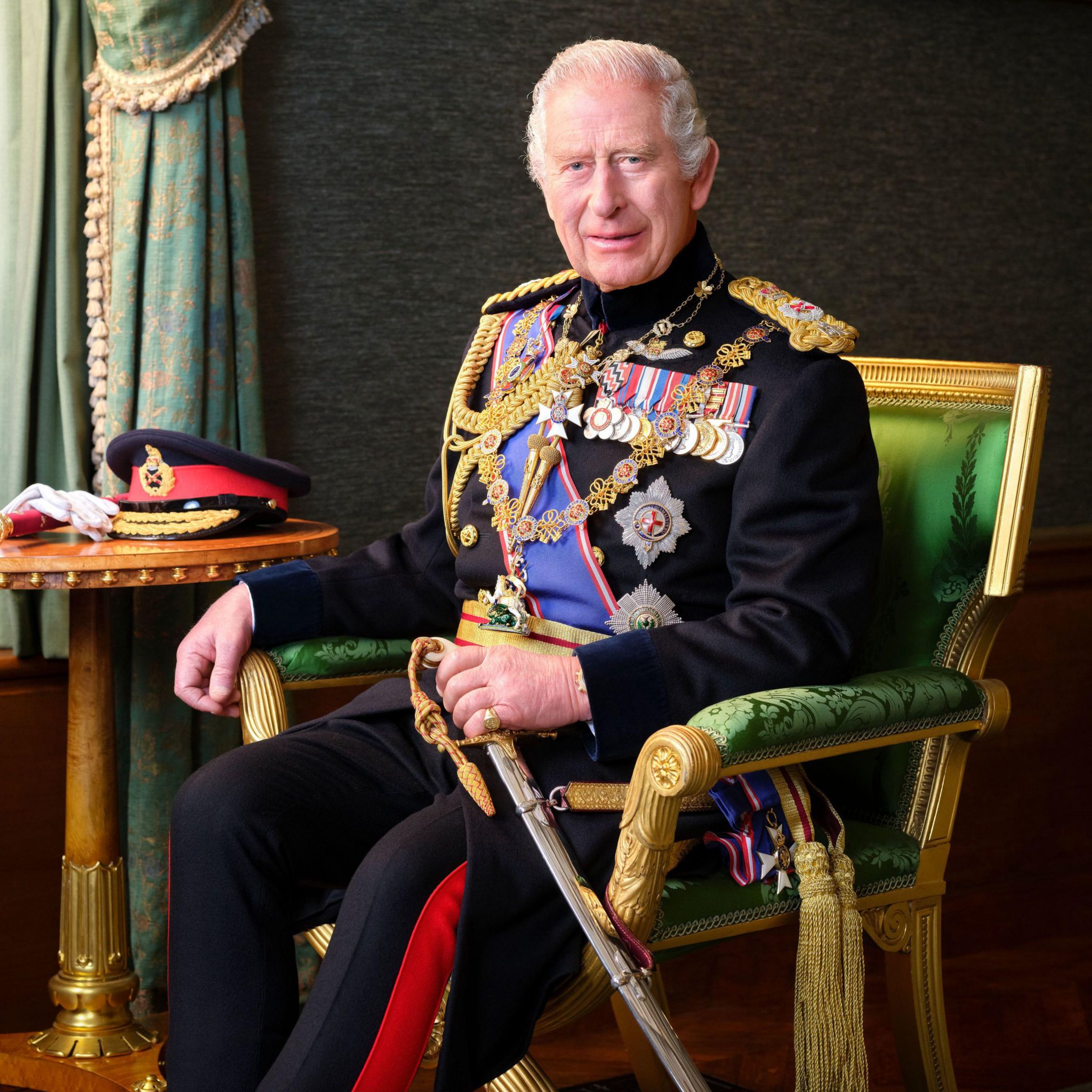 The photograph of King Charles, in which he is seated in ornate green and gold chair, wearing his full dress uniform with a ceremonial sword and ceremonial hat sitting on a table next to him