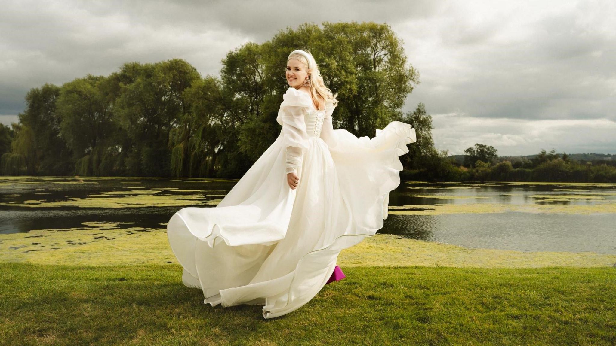 Brooke Jones is in a green field surrounded by ponds. She is wearing a white layered wedding dress and pink heels. She has blonde hair and a white headband. The dress is flowing in the wind