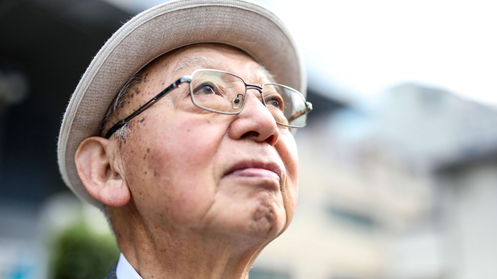 Portrait photo of Sueichi Kido, who is looking up and to the right of the camera. He is an elderly man with wire framed glasses and wearing a hat. The background is blurred.