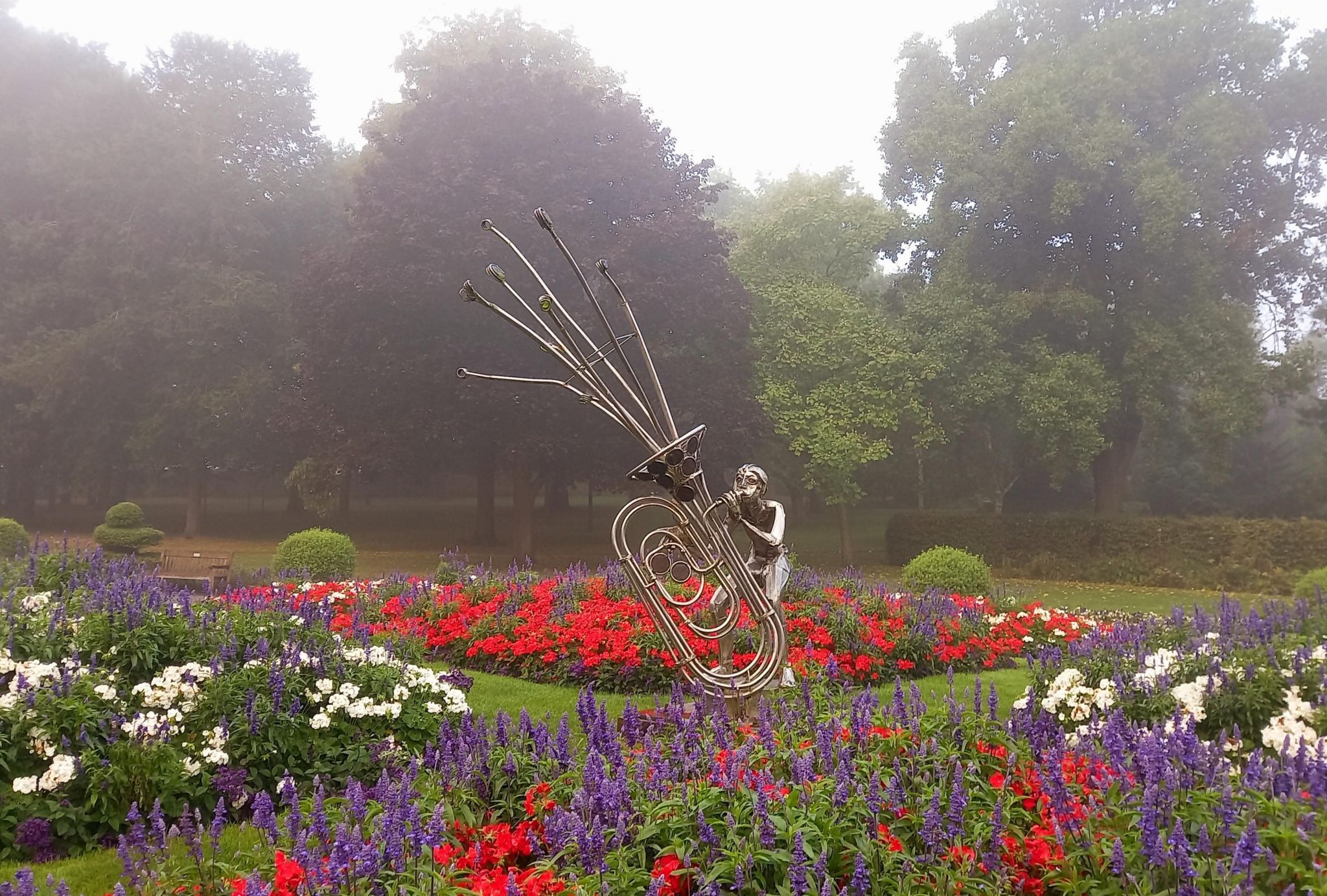 A metal sculpture of a child playing a tuba, standing in a park with beds of red, white and blue flowers around it.