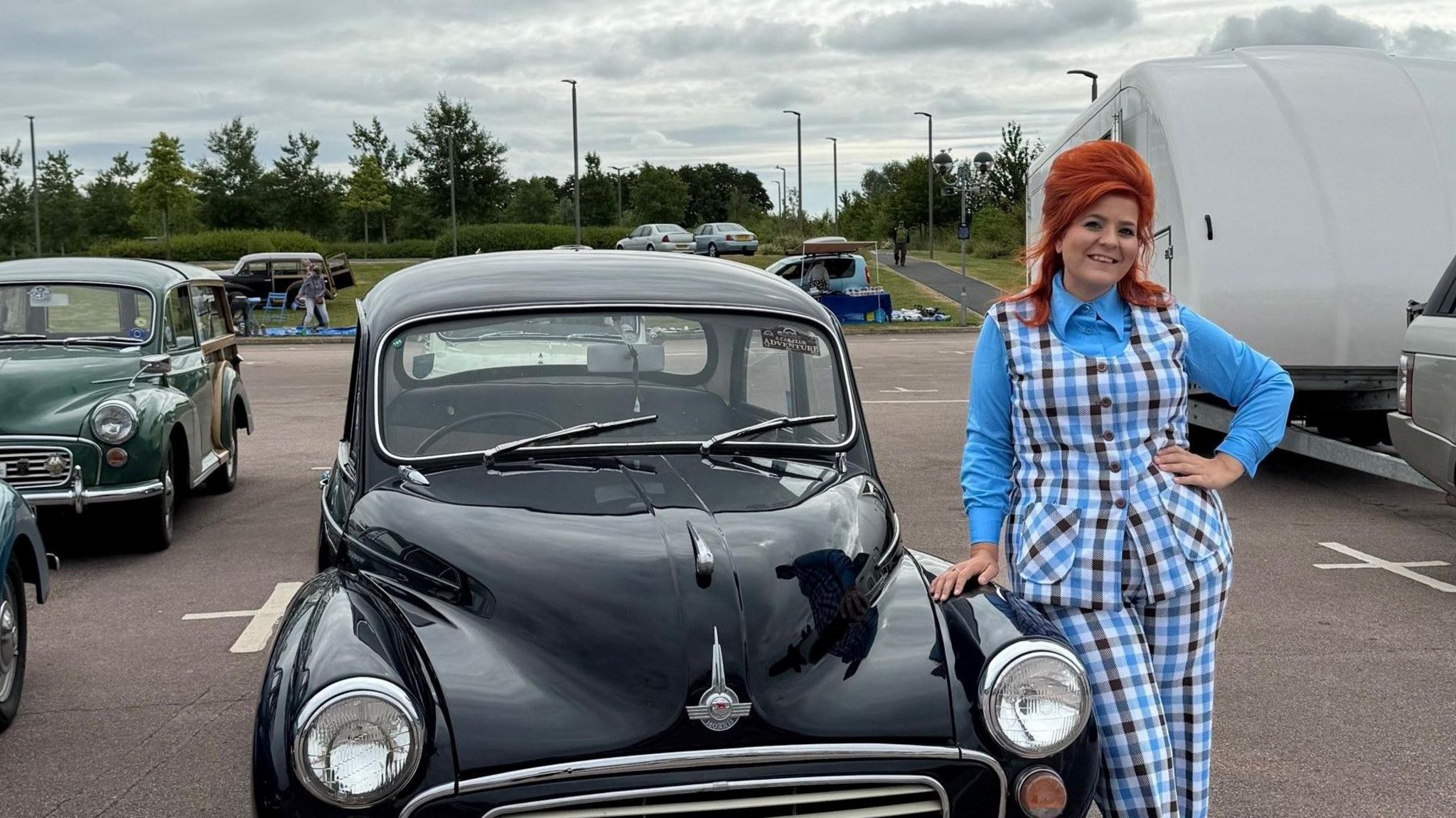 Steph Holloway leans against a black classic car, with her hand resting on the bonnet. She has red hair styled in a beehive and is a wearing a checked two-piece suit with a blue shirt