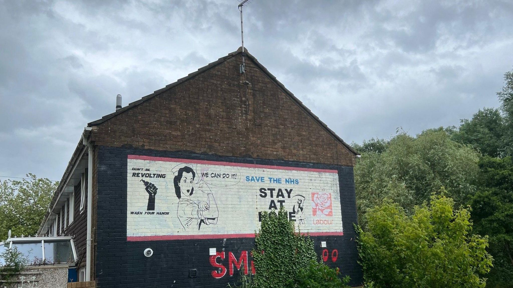 A mural depicting a nurse flexing her muscles with slogans saying "save the NHS" and "stay at home" on the end of a row of terrace housing