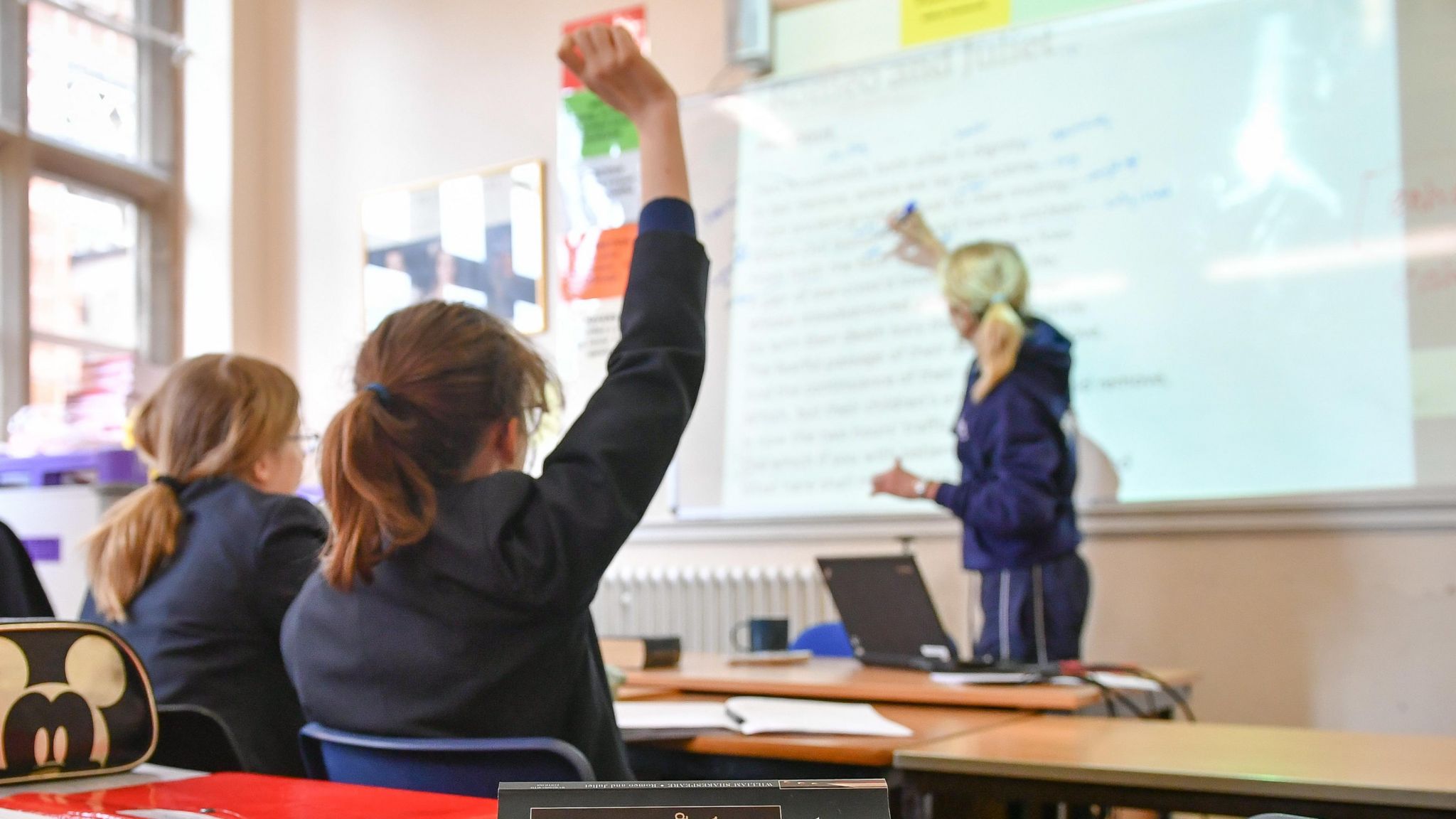 School children in classroom holding their hands up, next to an empty desk, while teacher writes on a white board