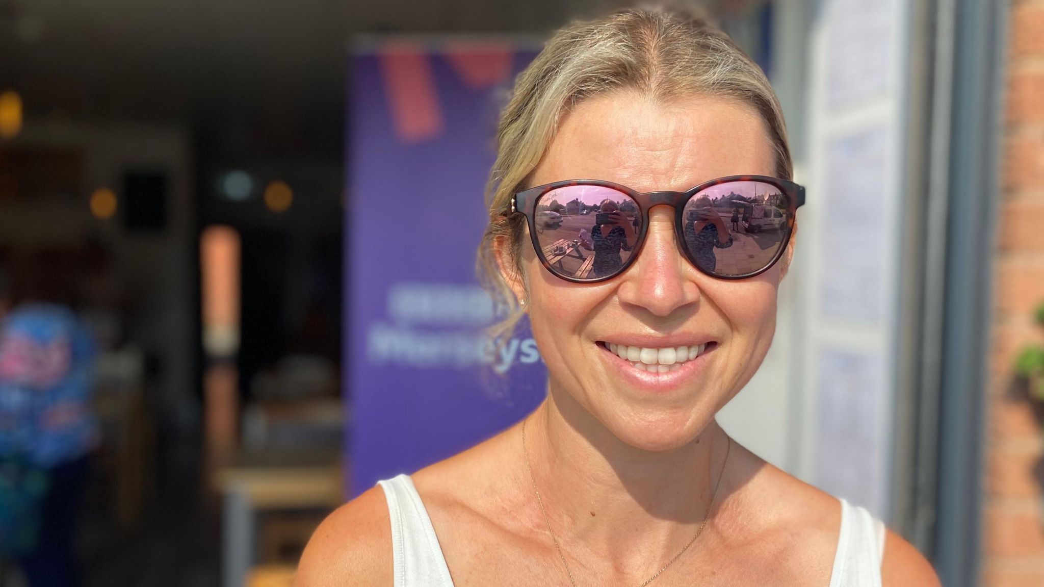 A smiling Rachel Lightburn, with blonde tied-back hair, reflective sunglasses and wearing a white vest top, stands in front of an open cafe door, with purple BBC Radio Merseyside branding behind her