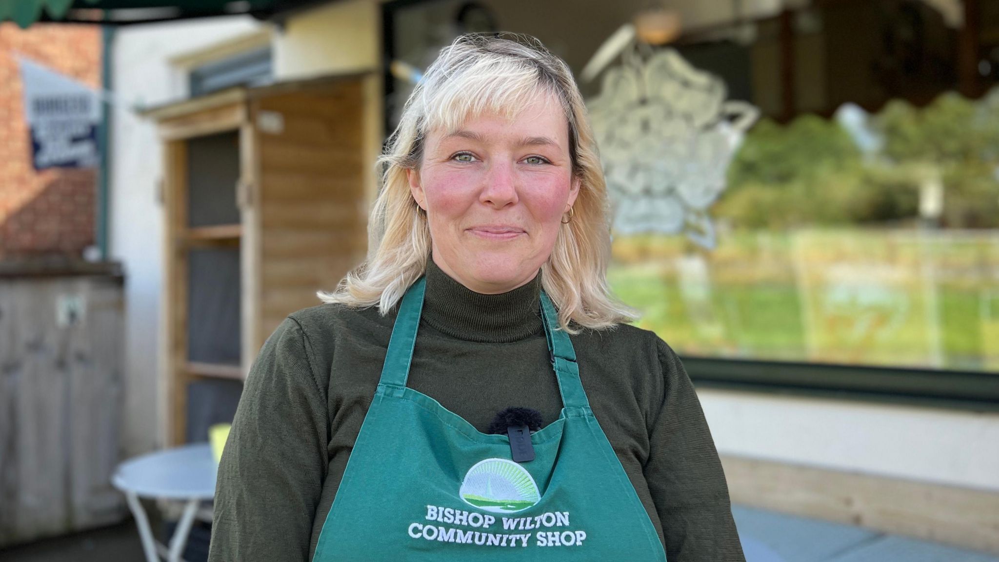 Annina Dyston has a long blonde bob and a fringe. She's wearing a green turtle neck jumper with an apron, which is a lighter shade of green, on top. She's standing in front of the Bishop Wilton Community Shop. 