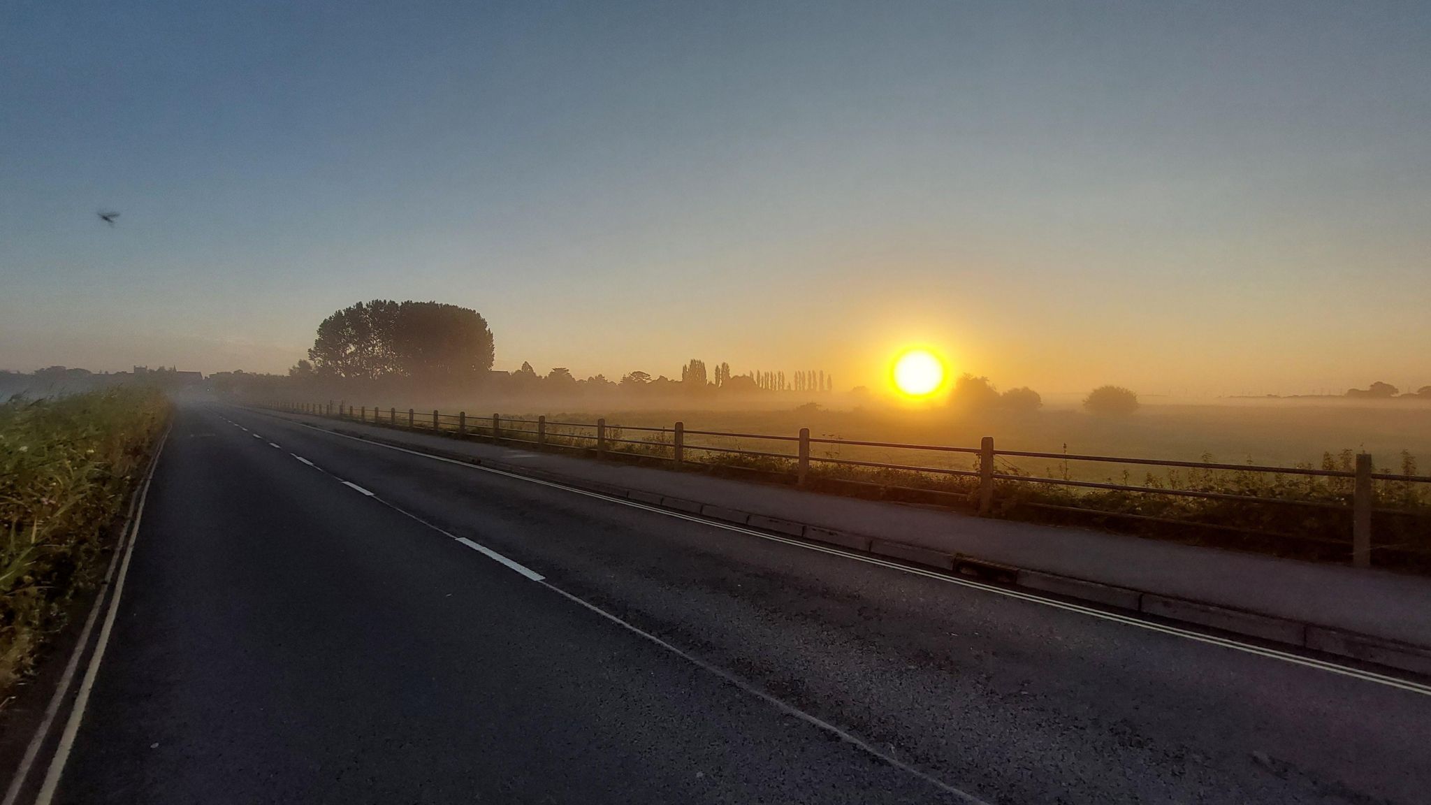 A road runs into the distance on the left handside of the frame with a field on the right separated by a low fence. A bright, yellow sun can be seen just above the horizon in the background.