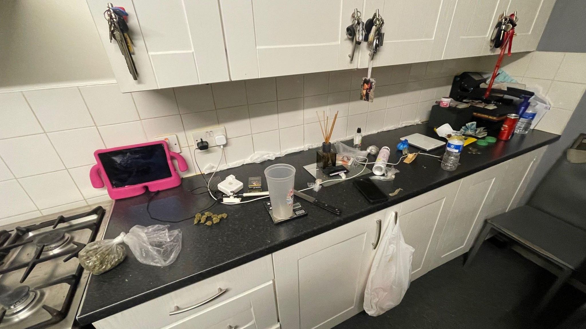 A messy kitchen worktop, which has a knife, drugs, money and other items cluttering it. The worktop is black and the cabinets are cream.