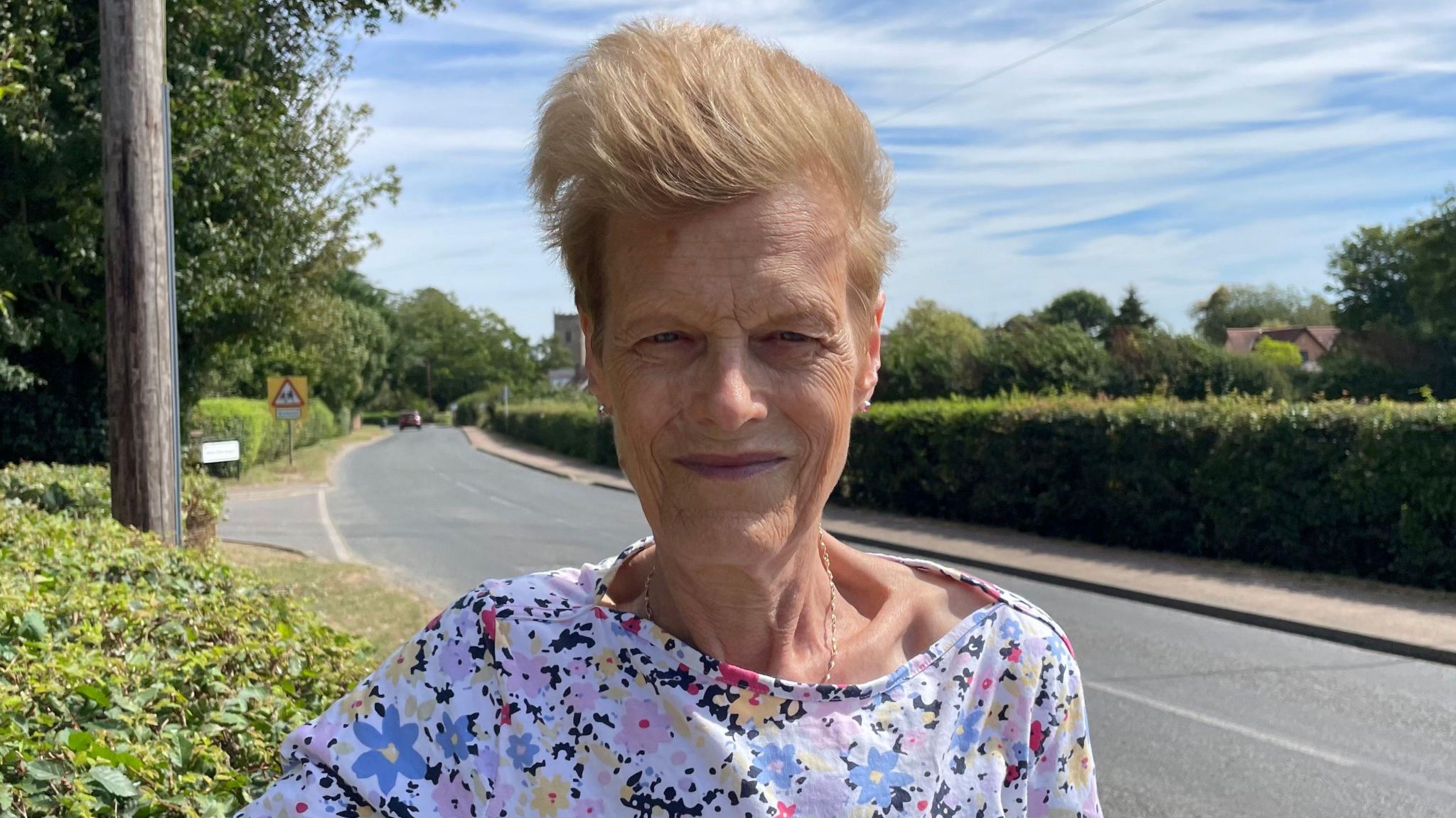 Looking direct to camera, Vanessa Kingsley is wearing a flowery top and standing by the roadside in Bacton. Her short, light brown hair is being swept back by a gust of wind and she is smiling gently. A mature thick hedge runs along the road in the background, bordering a narrow village road. A church is just visible in the far background.