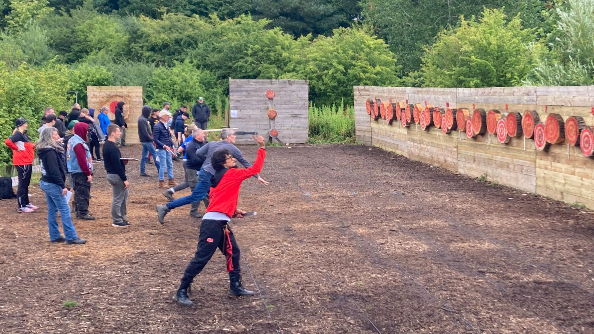 Two people throw axes towards round red targets fixed to a wooden wall, about five metres away. They are watched by about two-dozen spectators.