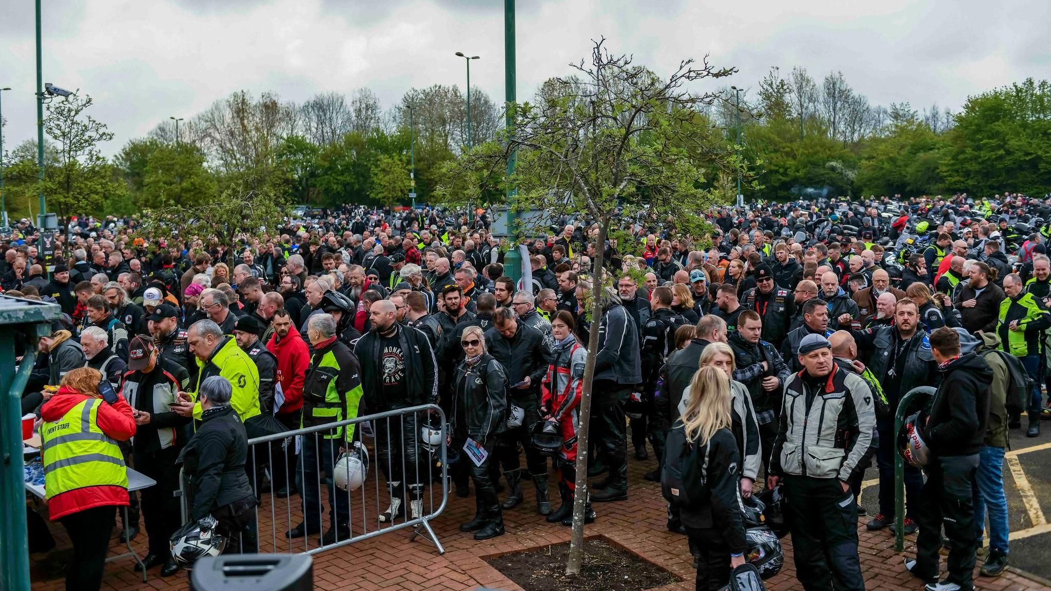 Thousands of people stand in a large crowd. There are grey metal barriers, and people in yellow hi-vis jackets