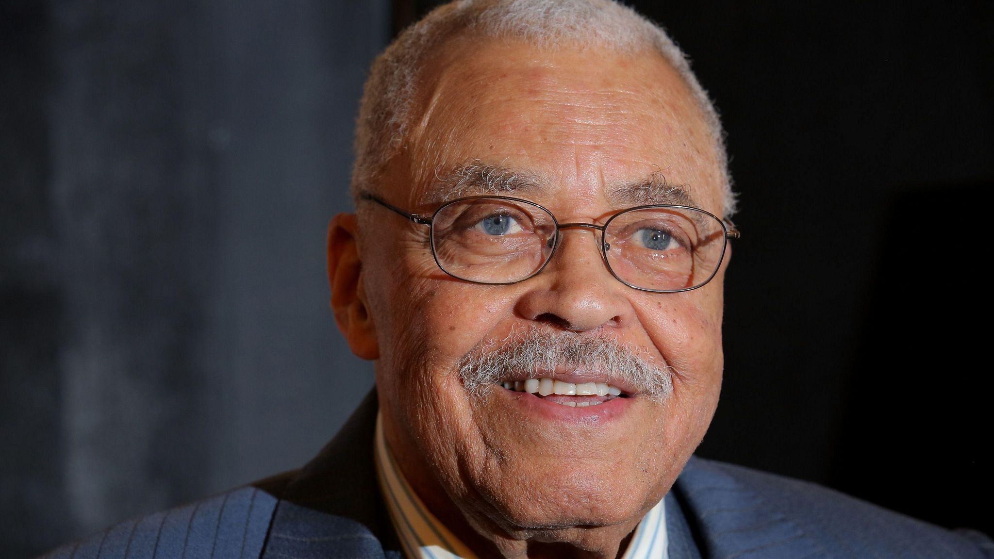 James Earl Jones wearing round spectacles looks towards the direction of a camera and smiling. The actor has a grey moustache and short grey hair. 