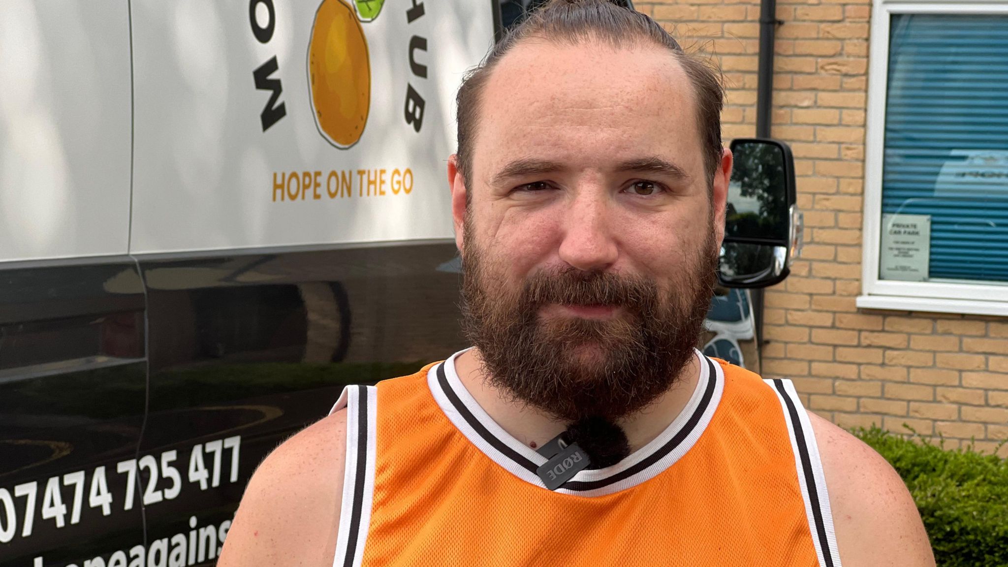 A man with dark beard and swept back dark hair wearing an orange vest standing in front of a van on his left and a wall on his right