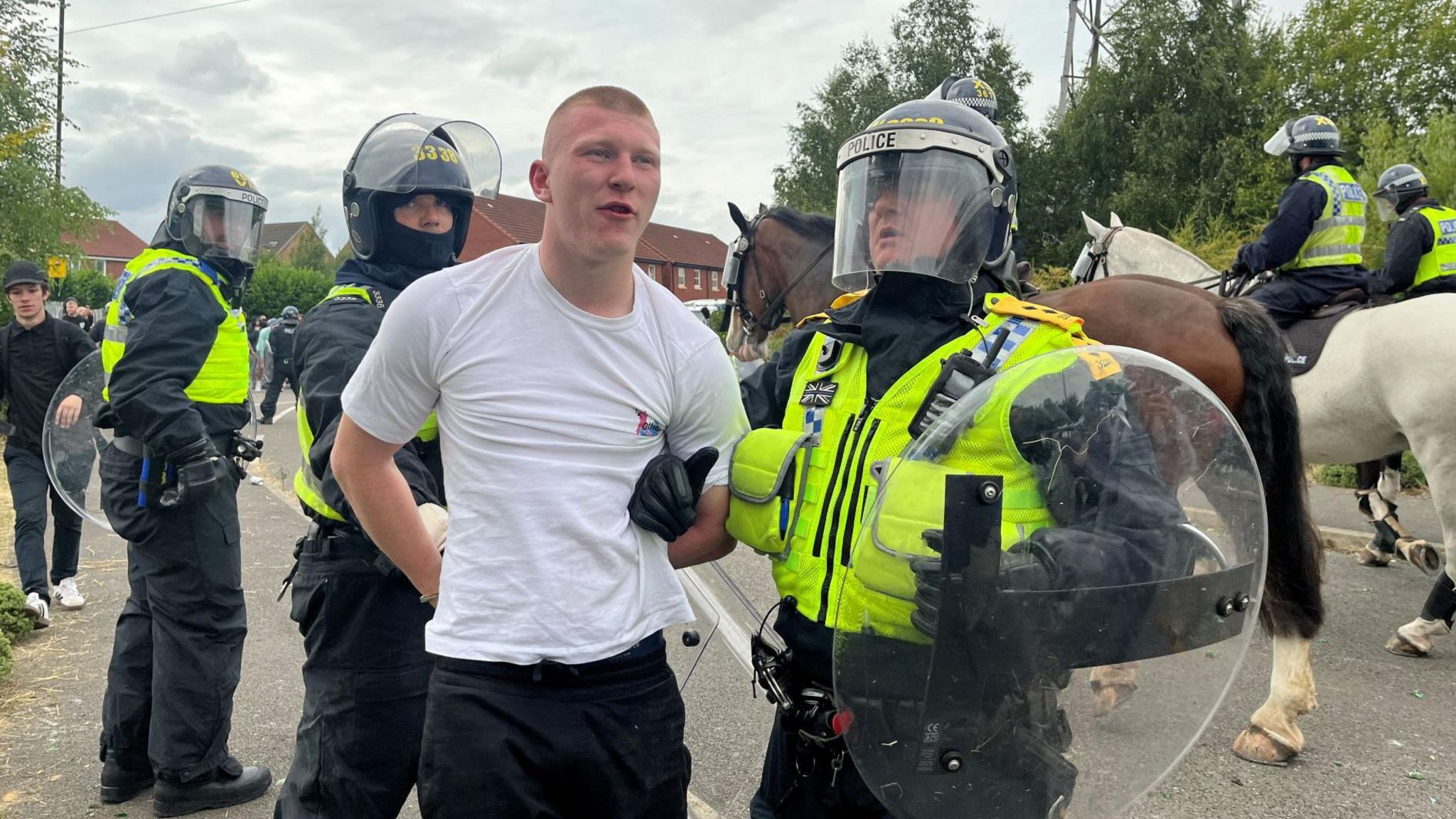 Gray is arrested by two officers who are wearing riot shields and helmets. A mounted officer can be seen in the background together with several other officers on foot. Gray is wearing a white t-shirt and black trousers. He has blond, shaven hair.