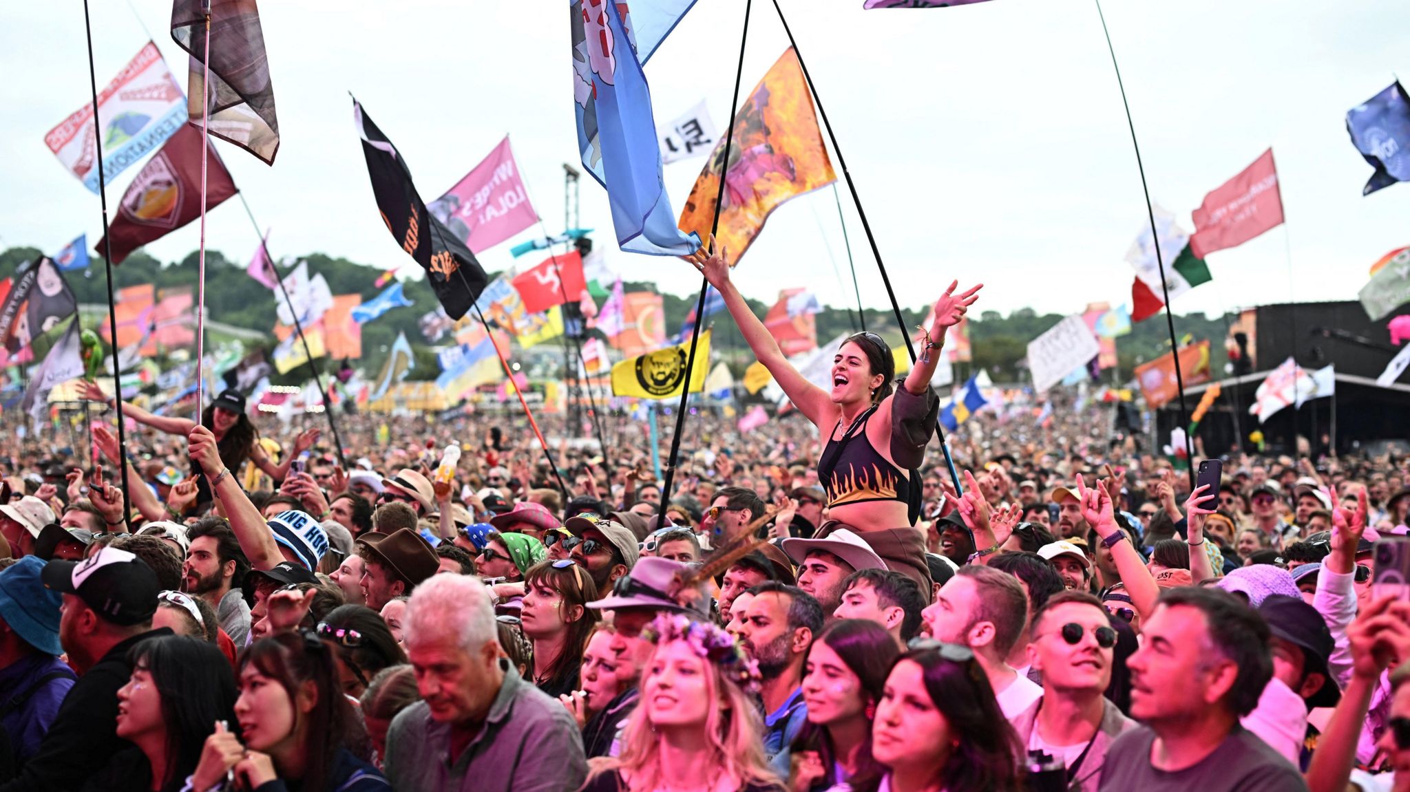 Image of the crowd at Glastonbury Festival. Flags can be seen, as well as one woman sat on somebody's shoulders with her hands in the air 