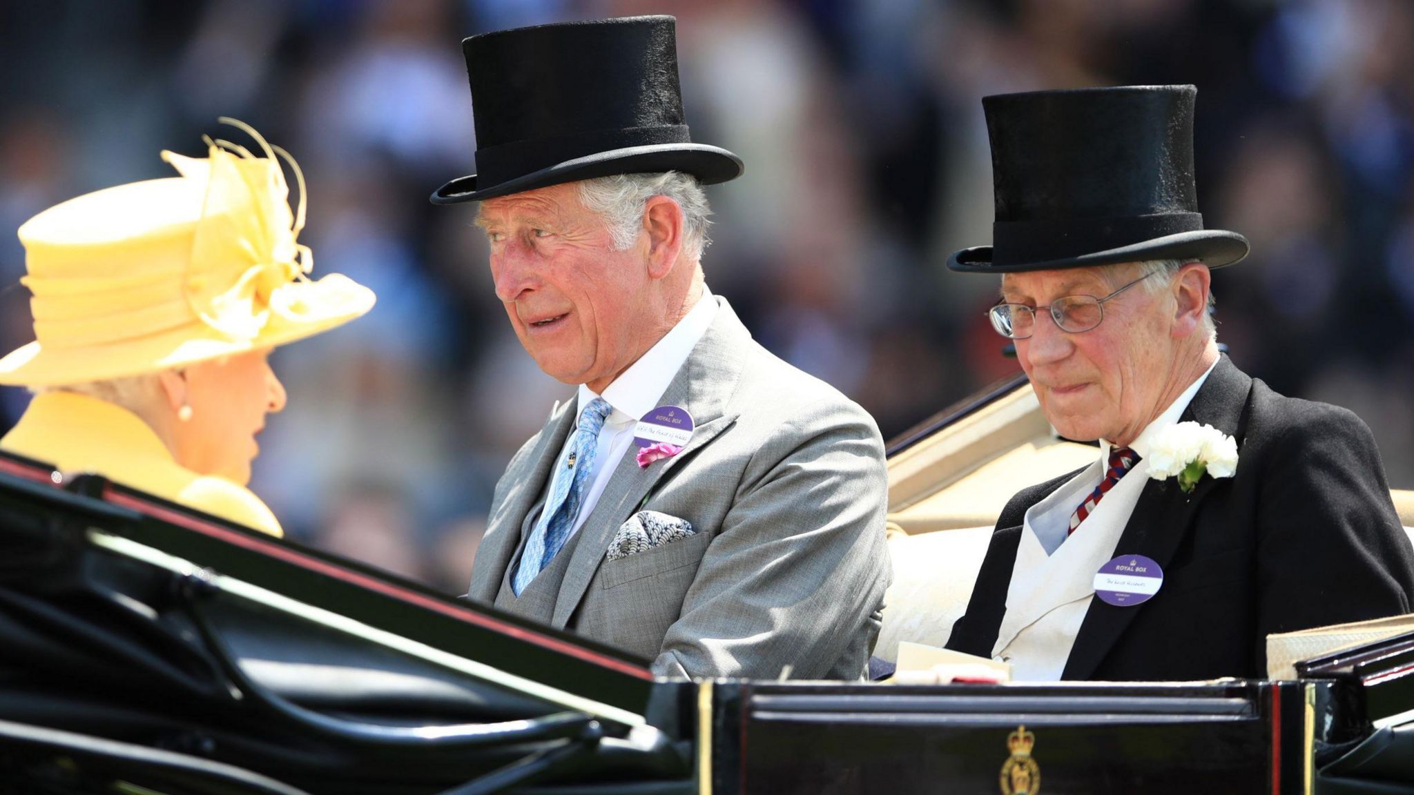 Queen Elizabeth II sits in an open carriage wearing a yellow hat. Lord Fellowes and the then-Prince Charles sit opposite her in suits and wearing black top hats