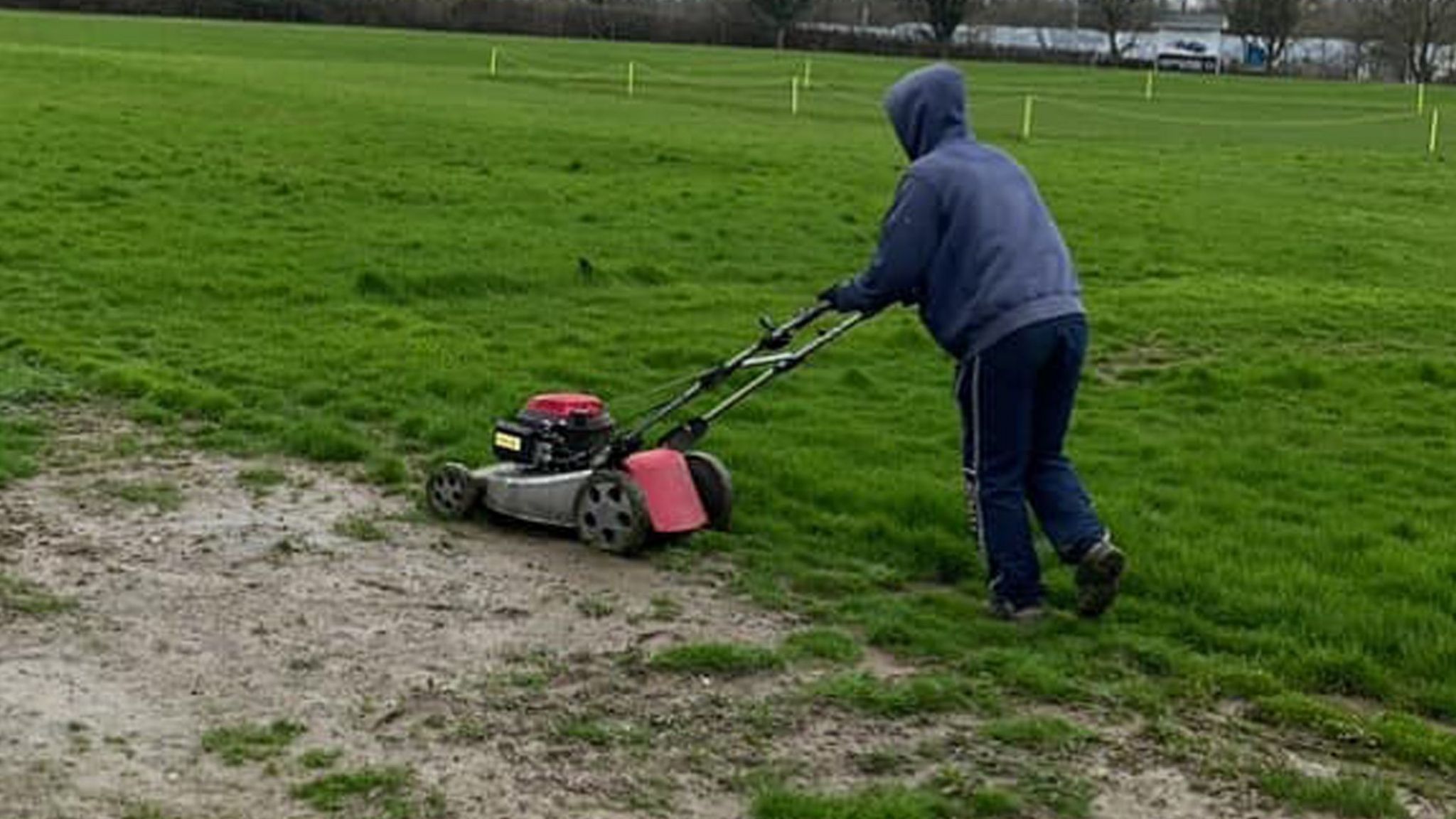 A man in a blue hoodie pushes a mower on the outfield, which is partly grassy but with a large muddy bald patch