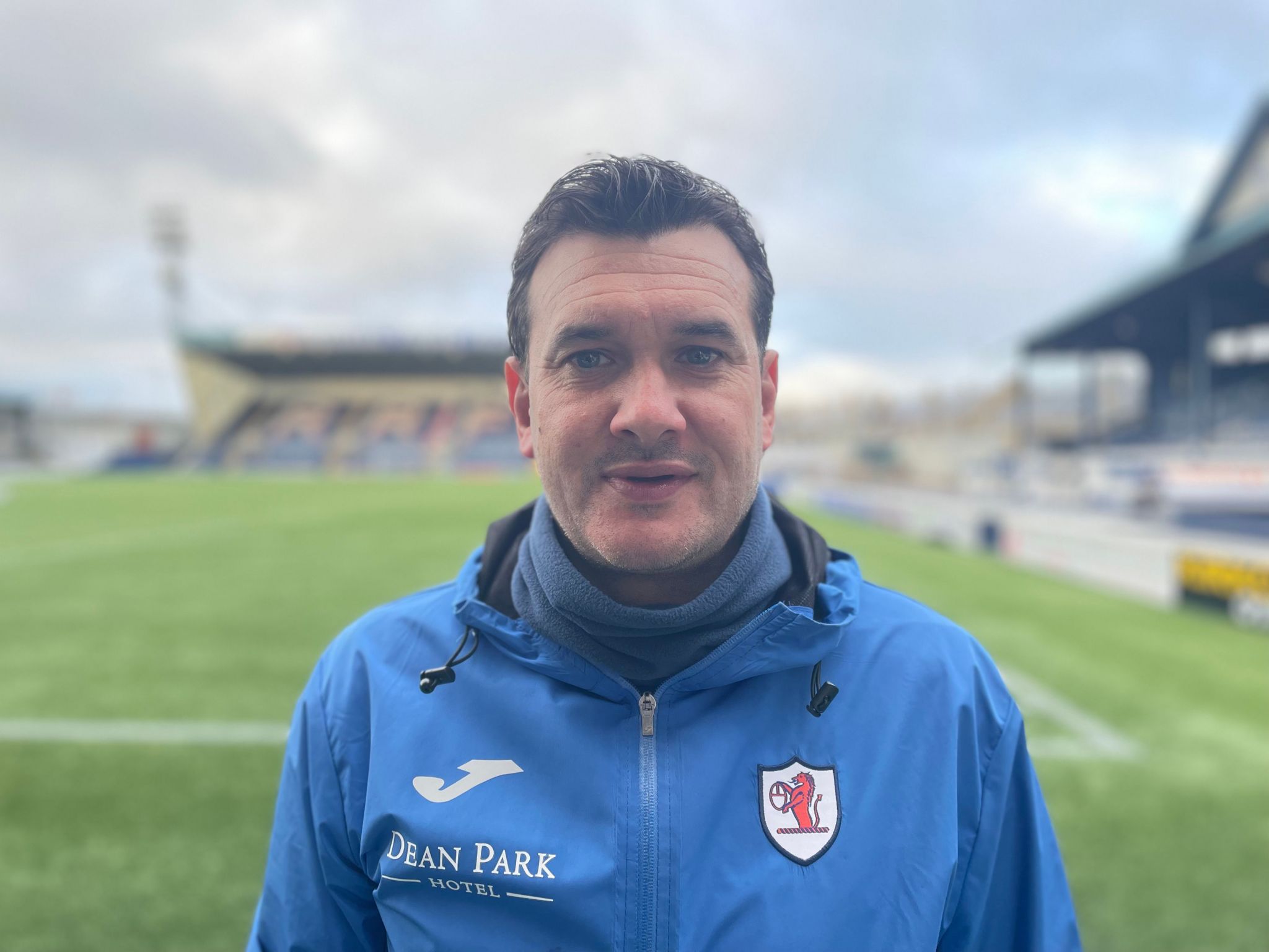 Raith manager, Ian Murray - man looking at camera, wearing a blue jacket with the club logo with a football pitch in the background out of focus.