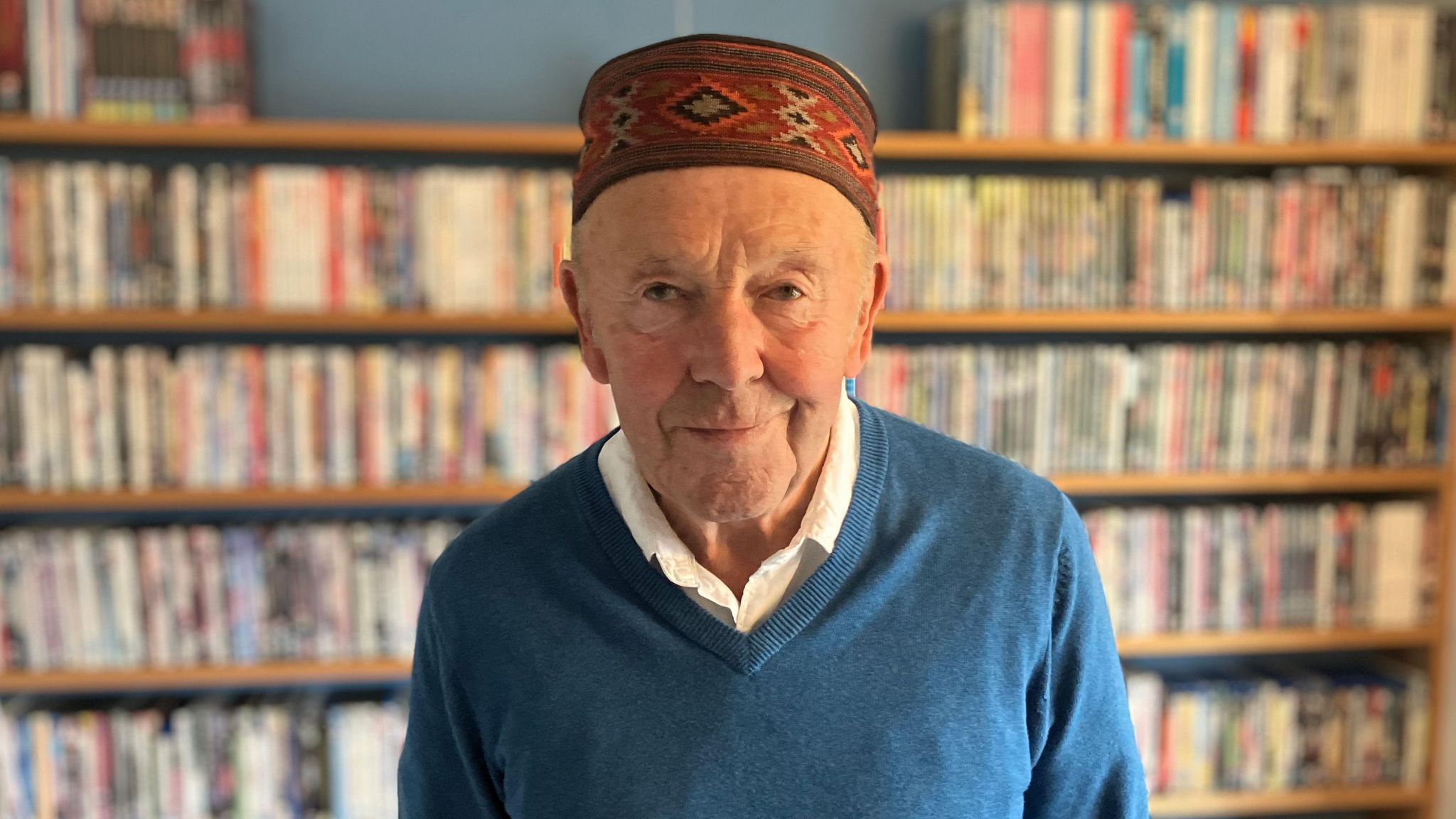 Mark Patterson, who is standing in front of a book shelf at his house. He is wearing a blue jumper with a white shirt underneath. He also has a red patterned hat on his head. He is smiling.