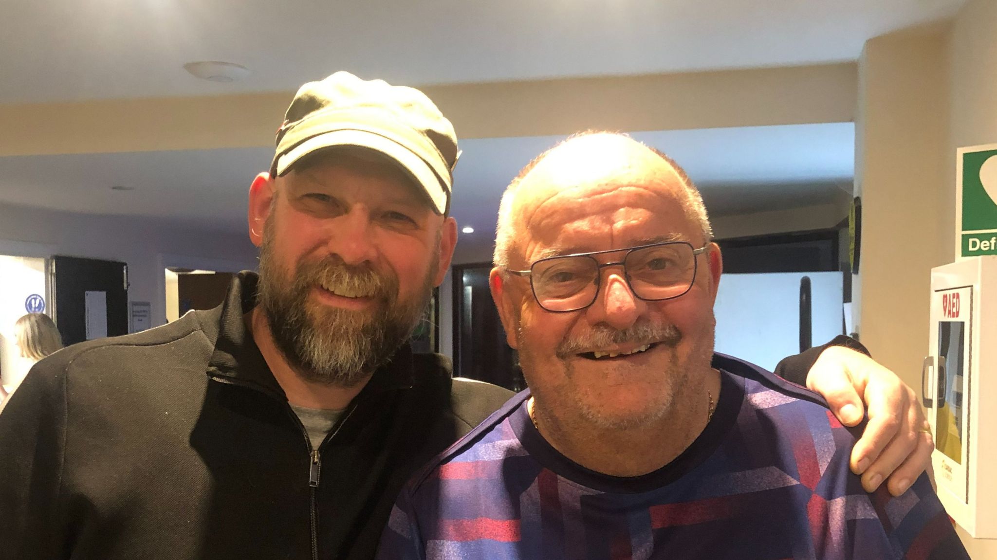 Man with beard and baseball cap with arm round man wearing glasses