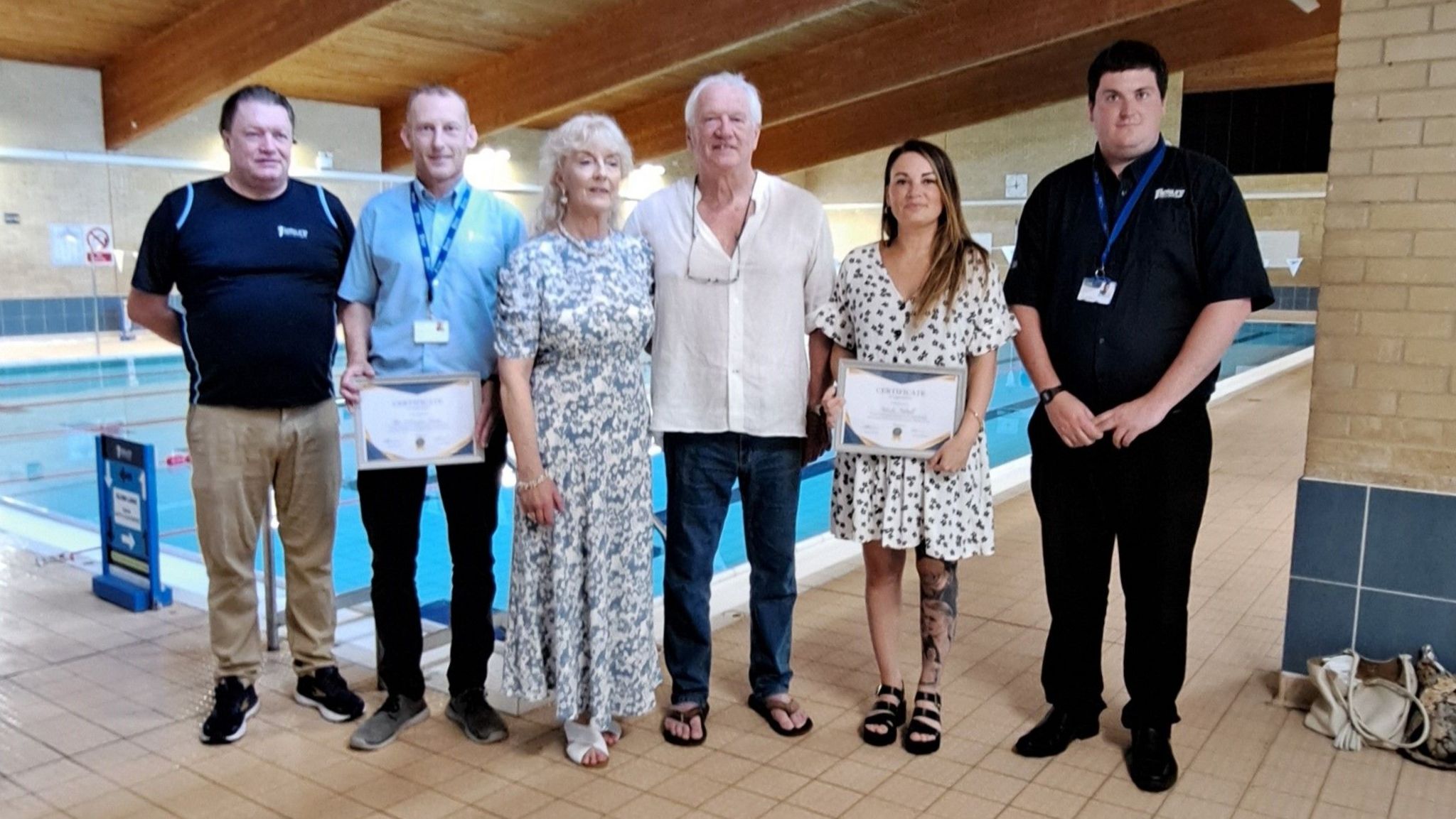 Two women and four men stand beside an indoor swimming pool. They are fully-clothed. One is holding a certificate.