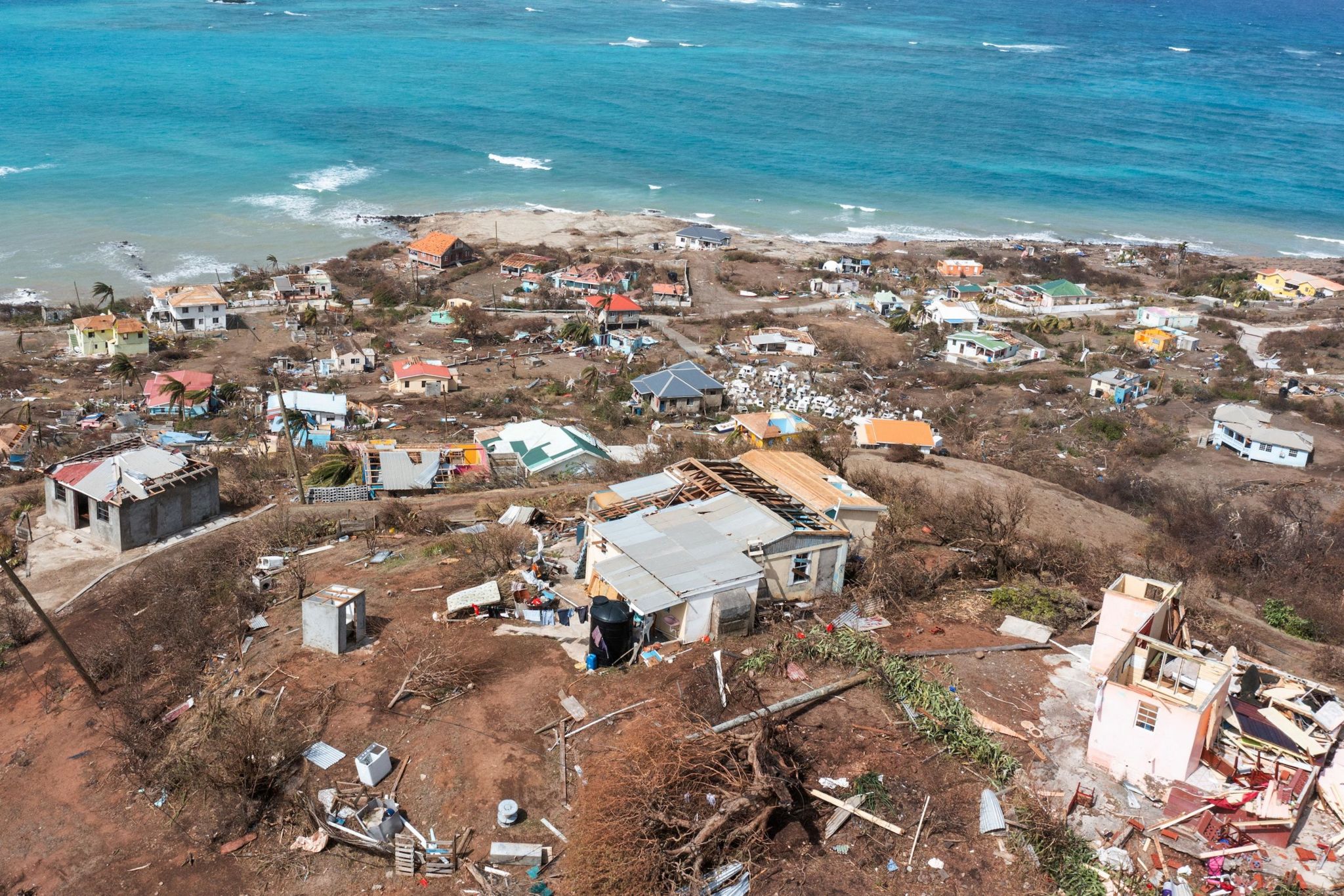 A birds-eye view of a hillside shows smashed houses leading to the sea