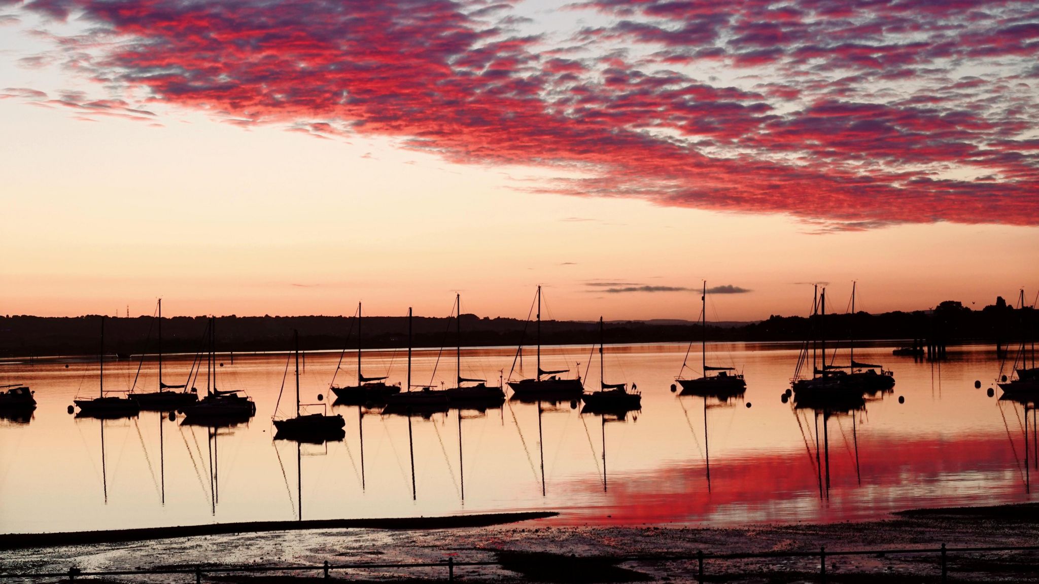 WEDNESDAY - The sunrise over Gosport with 14 small yachts silhouetted in the water in the foreground. The water in the harbour is glowing orange, reflecting the vivid red clouds above. On the horizon there are buildings and trees in silhouette against the orange sky.