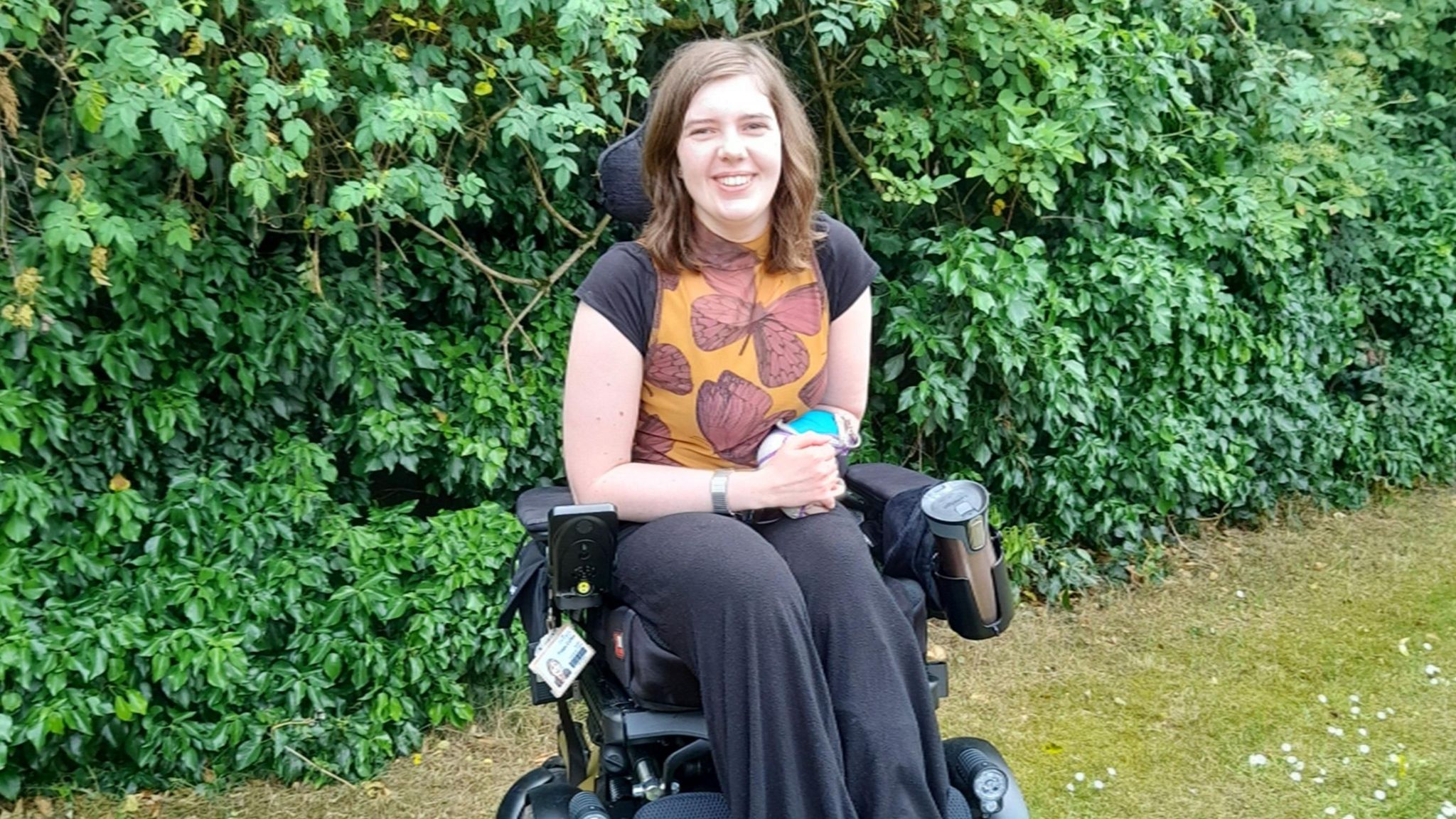 Poppy Garton pictured in a wheelchair and smiling at the camera. Poppy is wearing an orange top with butterflies on and black trousers. 