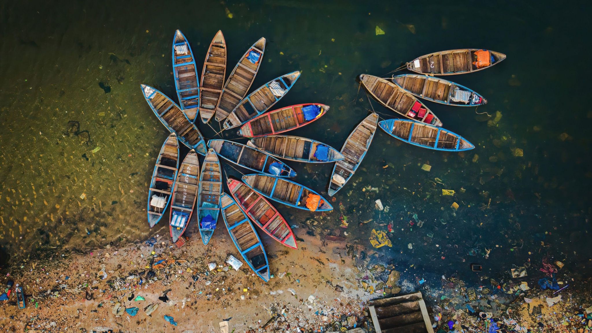 Alex Cao's photo 'Boat flower in pollution', which shows an aerial view of a number of wooden boats tethered together and floating so they resemble petals. The water and beach is shown covered in pollution. 