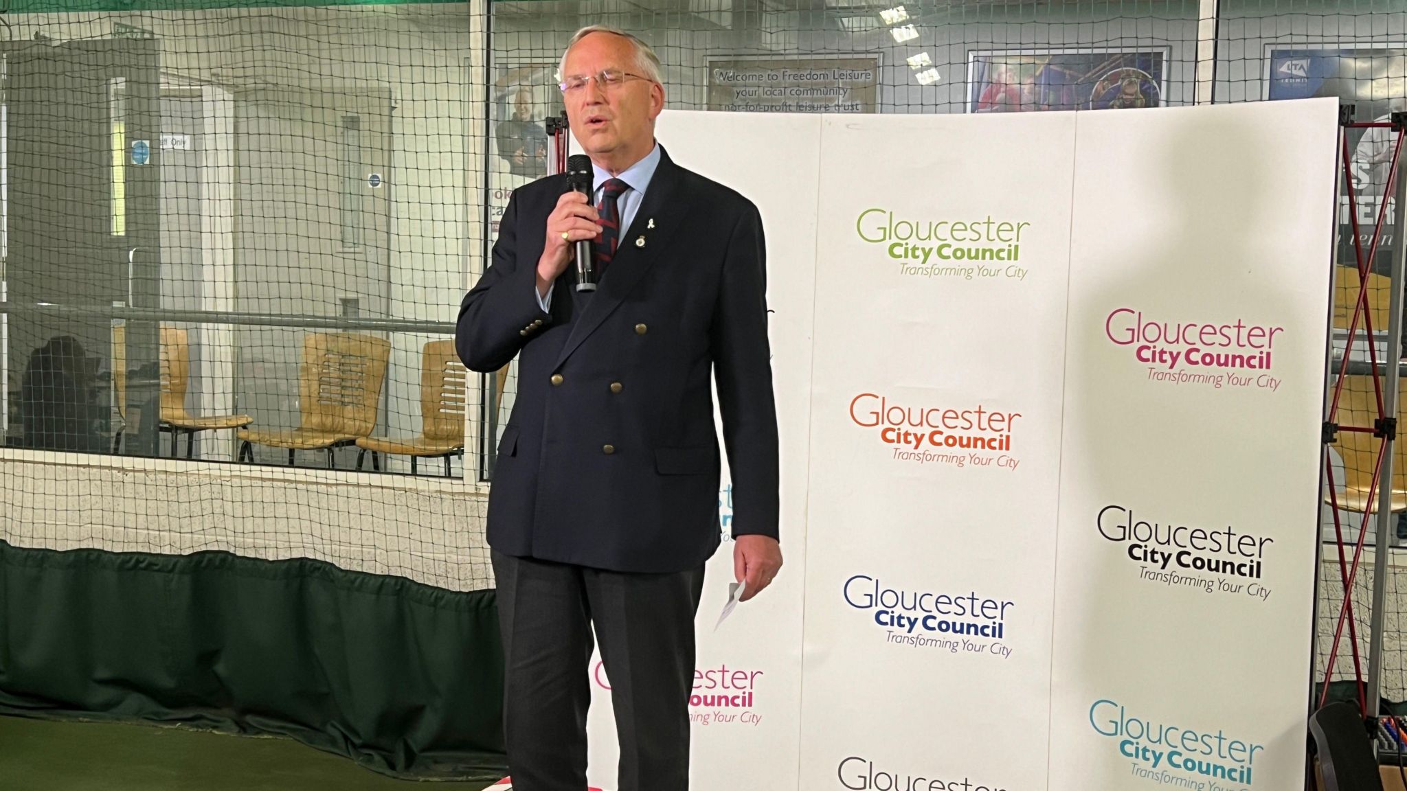 Chris Nelson stands in front of a white background branded with Gloucester City Council written in different colours. He is holding a microphone and wearing a dark blue-black suit buttoned up