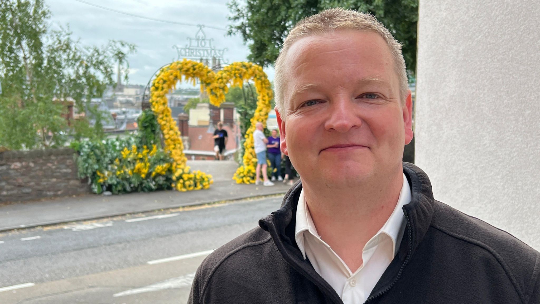 Chris Pitt wearing a white collared shirt and black three quarter zip sweatshirt. He is standing across the road from the installation, which is visible in the background