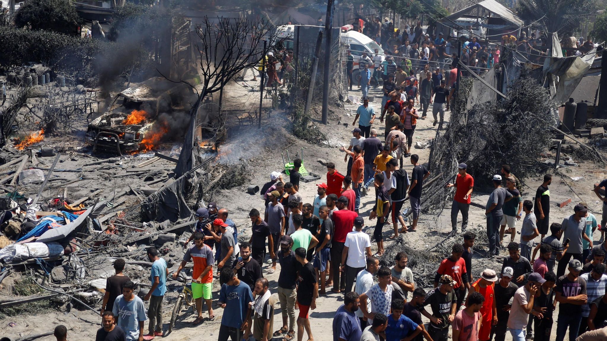 People look at the aftermath of an Israeli airstrike in Gaza. A burning car is visible to the left of the image.