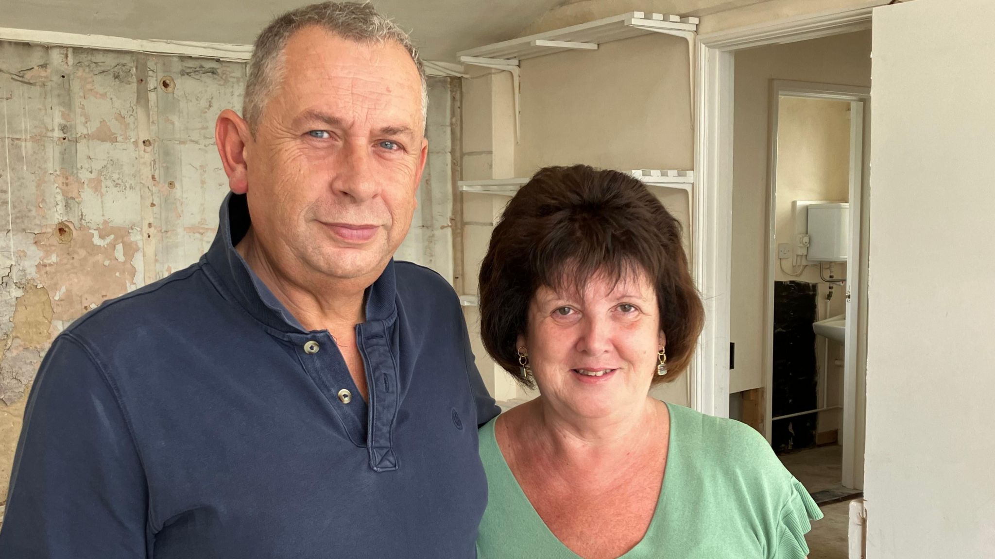 Dave and Donna Hammond standing in their store. Mr Hammond who has cropped hair is wearing a blue polo shirt. Mrs Hammond is shorter, with bobbed brown hair, and wearing a green T-shirt. The walls of the shop in the background are bare and show water damage. 