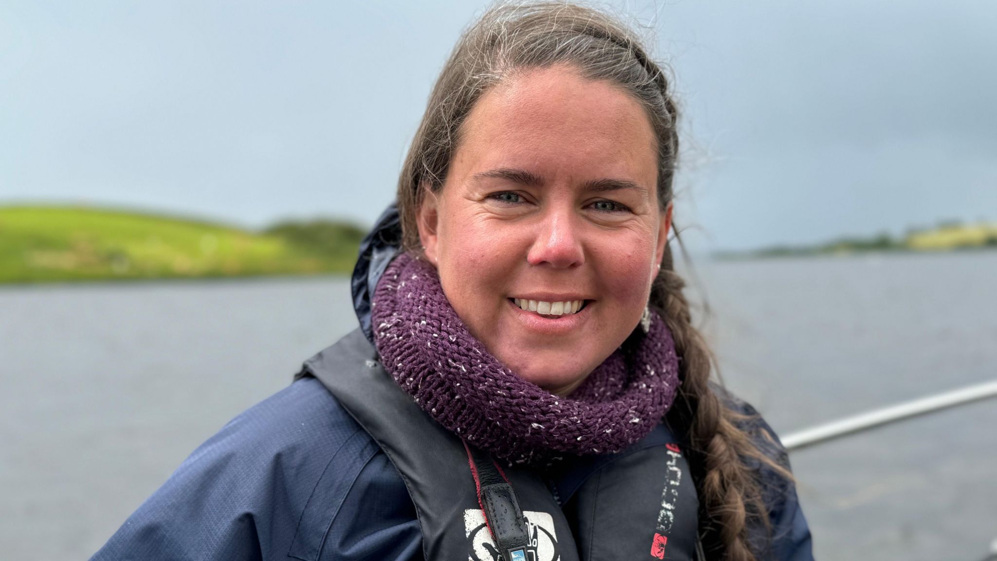 Headshot of Amy Burns smiling into the camera. She is wearing a life-jacket. In the background a body of water and beyond that a grassy hill.