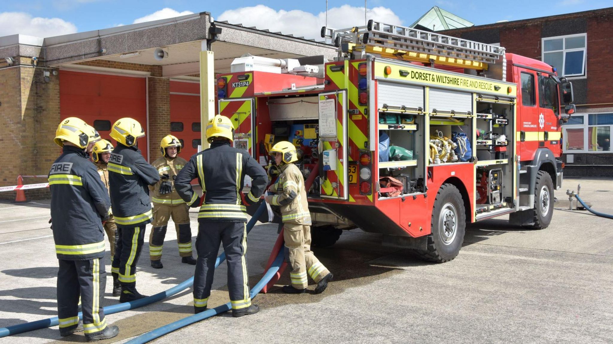 A crew of five firefighters in uniform standing outside a fire engine. The vehicle is parked at a fire station. There are two blue hosepipes coming from the back of the fire engine.