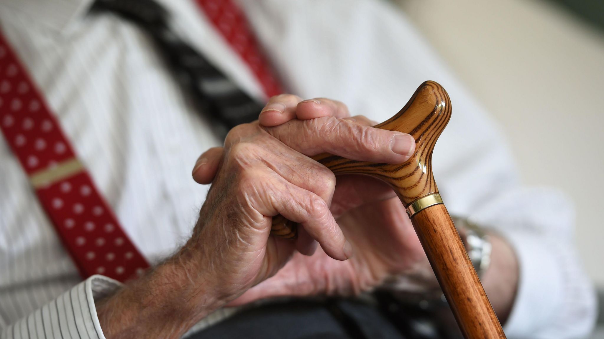 Close up of an elderly person's hands holding onto a walking stick, wearing a pinstriped shirt with red braces and a tie