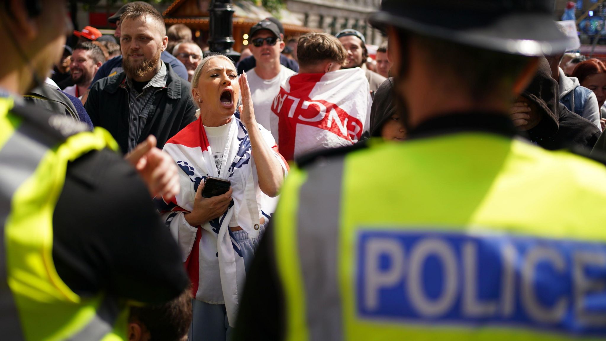 A woman, draped in an England flag, shouts at police while surrounded by a number of other male protesters, some also wearing England flags around their shoulders