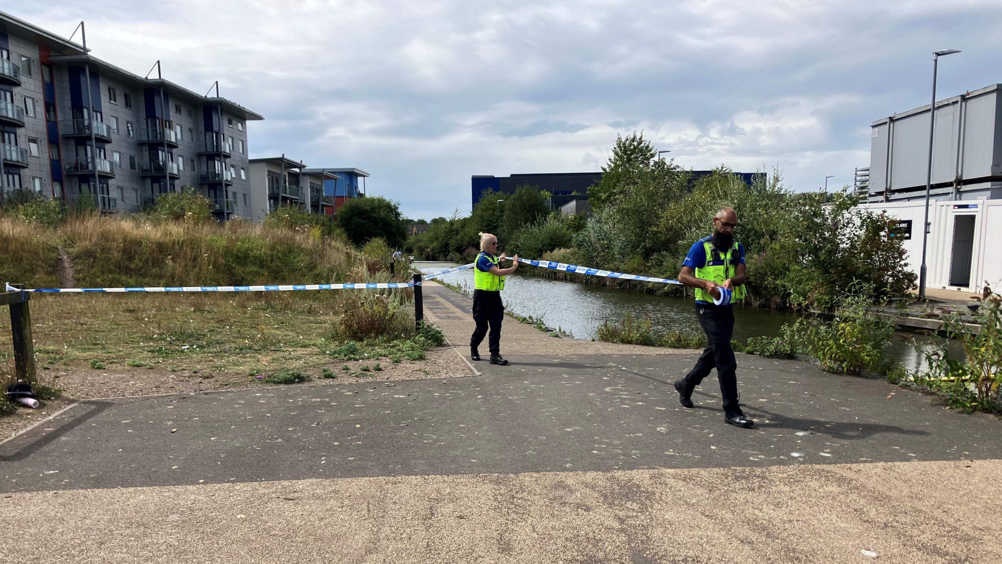 Two police officers unroll police tape at the scene, one is male with a black beard and the other female with blonde hair. They have attached the tape to a post next to a path by the canal 