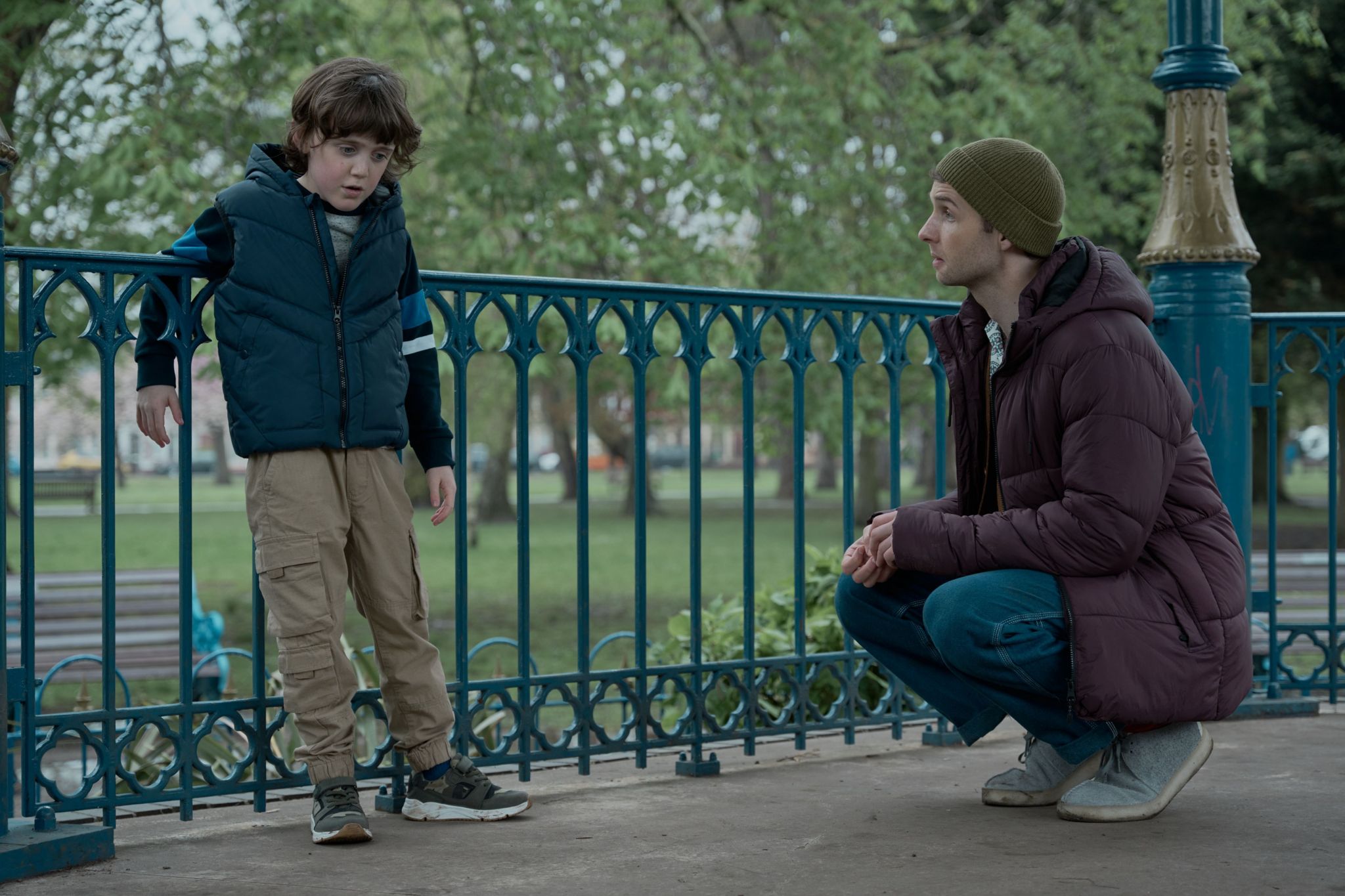 A young boy is leaning on a railing, as a man is bent down trying to speak with him.