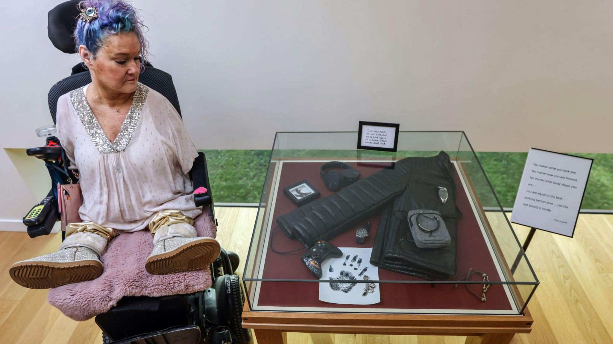 Alison Lapper looks at a collection of her son's belongings on a table next to her. The belongings include his coat, bag and games console. Alison is wearing a pink top and has purple dyed hair.