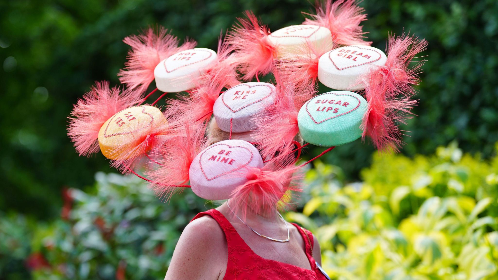 A woman wearing a hat, so large it obscures her face, with seven large attachments made to look like Love Heart Sweets