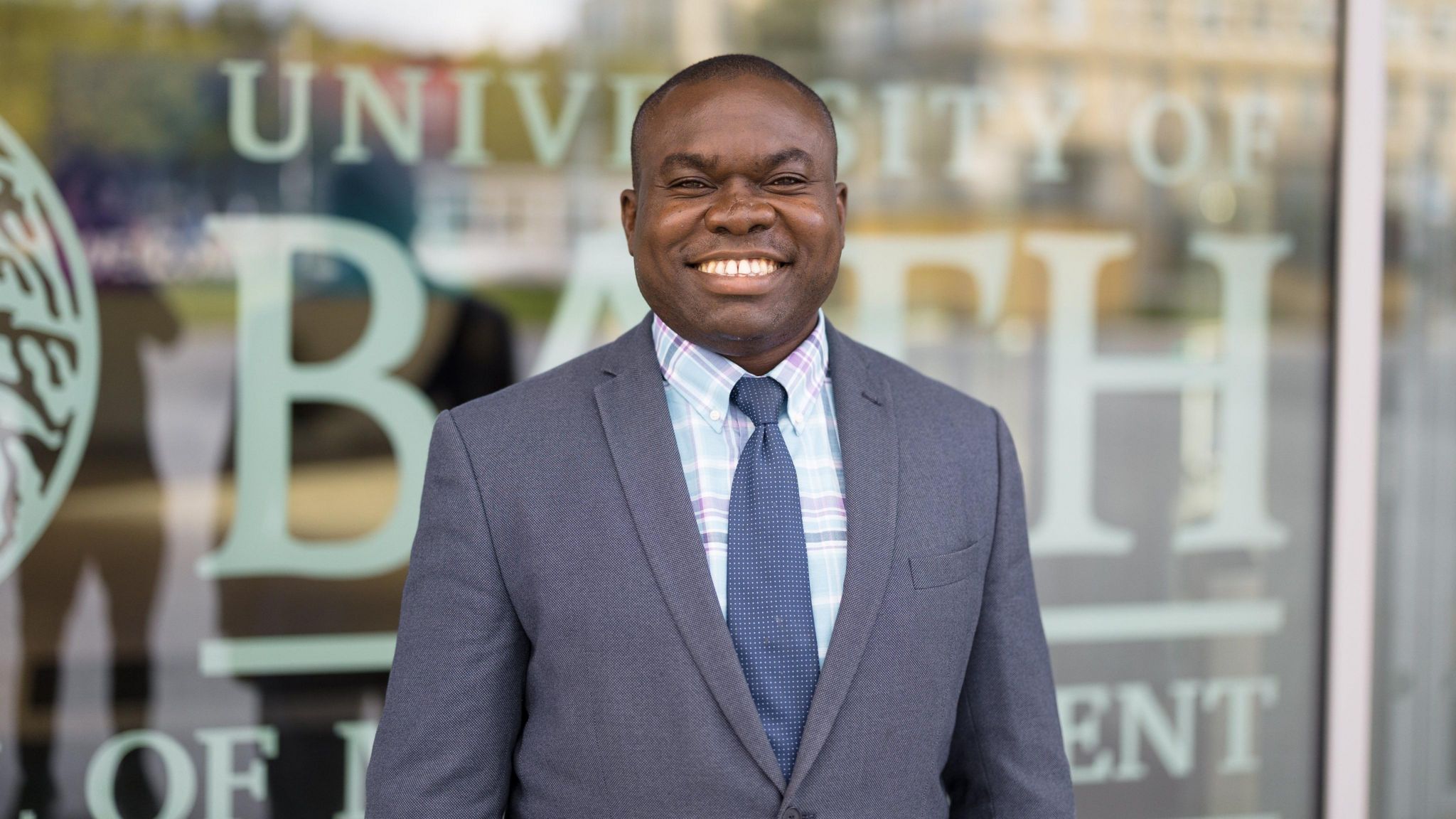 Dr Teslim Bukoye smiling at the camera wearing a dark grey suit in front of a window with University of Bath written on it