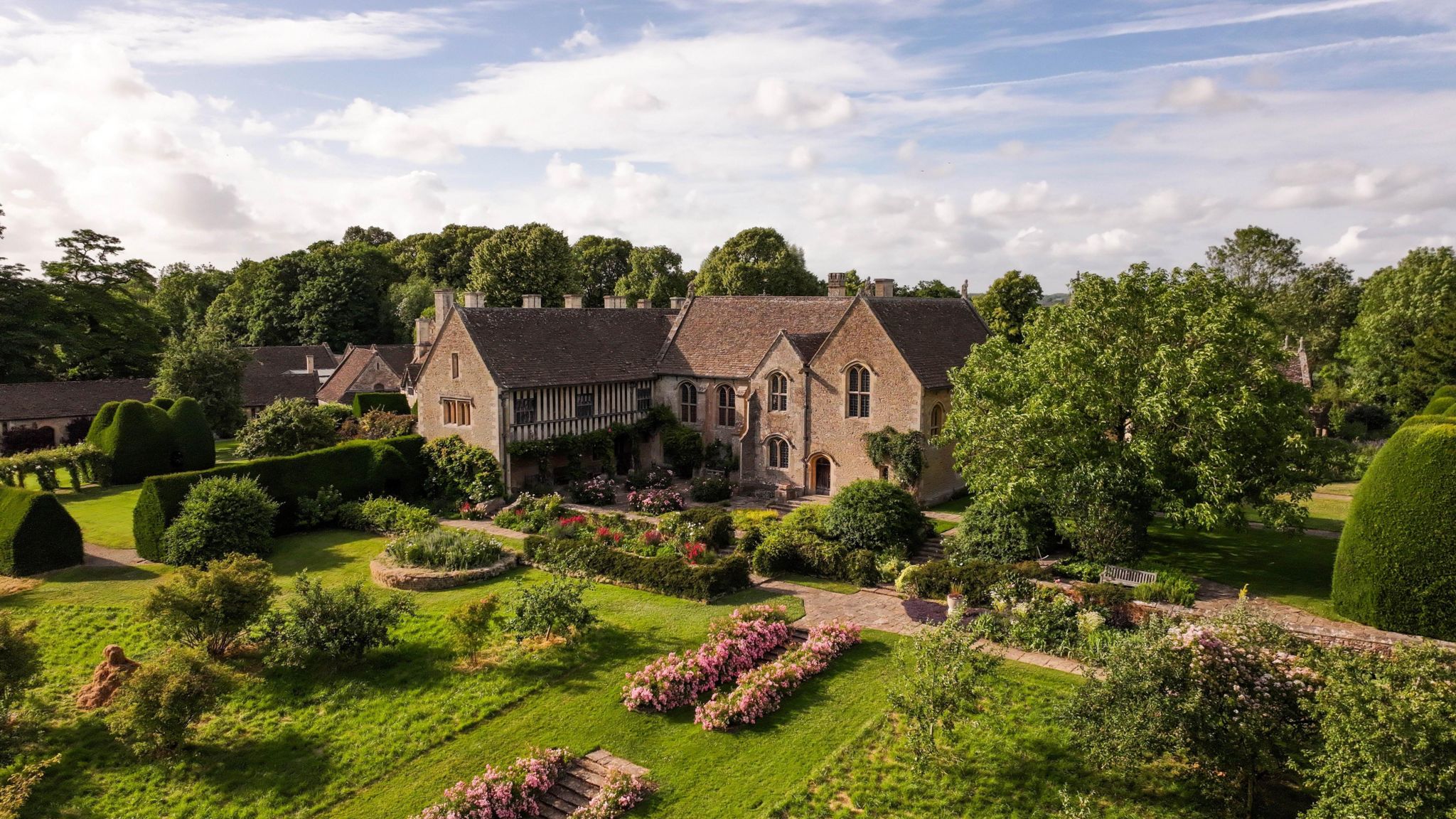 A brown Great Chalfield Manor with surrounding green gardens containing trees and flowerbeds on a sunny day seen from an aerial shot